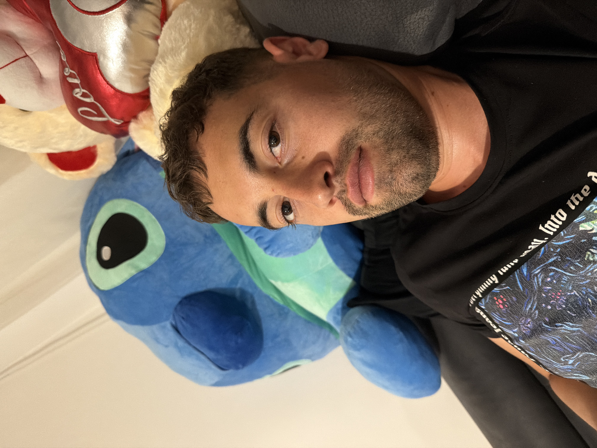 An indoor portrait captures a young man lying down, looking directly into the camera. He has short, curly dark hair, stubble, and brown eyes, with a neutral expression. He is wearing a black t-shirt featuring a colorful, intricate graphic pattern on the right sleeve, where partial white text reading "into the d..." is visible.
The man is positioned on a dark-colored surface, likely a sofa or bed. He is partially resting against a large, blue plush toy resembling Disney's Stitch. In the upper left corner of the frame, another plushie is visible; it is white and fluffy, with a prominent red, shiny section on which the word "Love" is written in white script. The background consists of a plain, light-colored wall towards the bottom left of the image. The image is rotated counter-clockwise, suggesting the perspective of someone lying down or a sideways phone orientation. The casual setting indicates a home environment located in Plovdiv, Bulgaria.