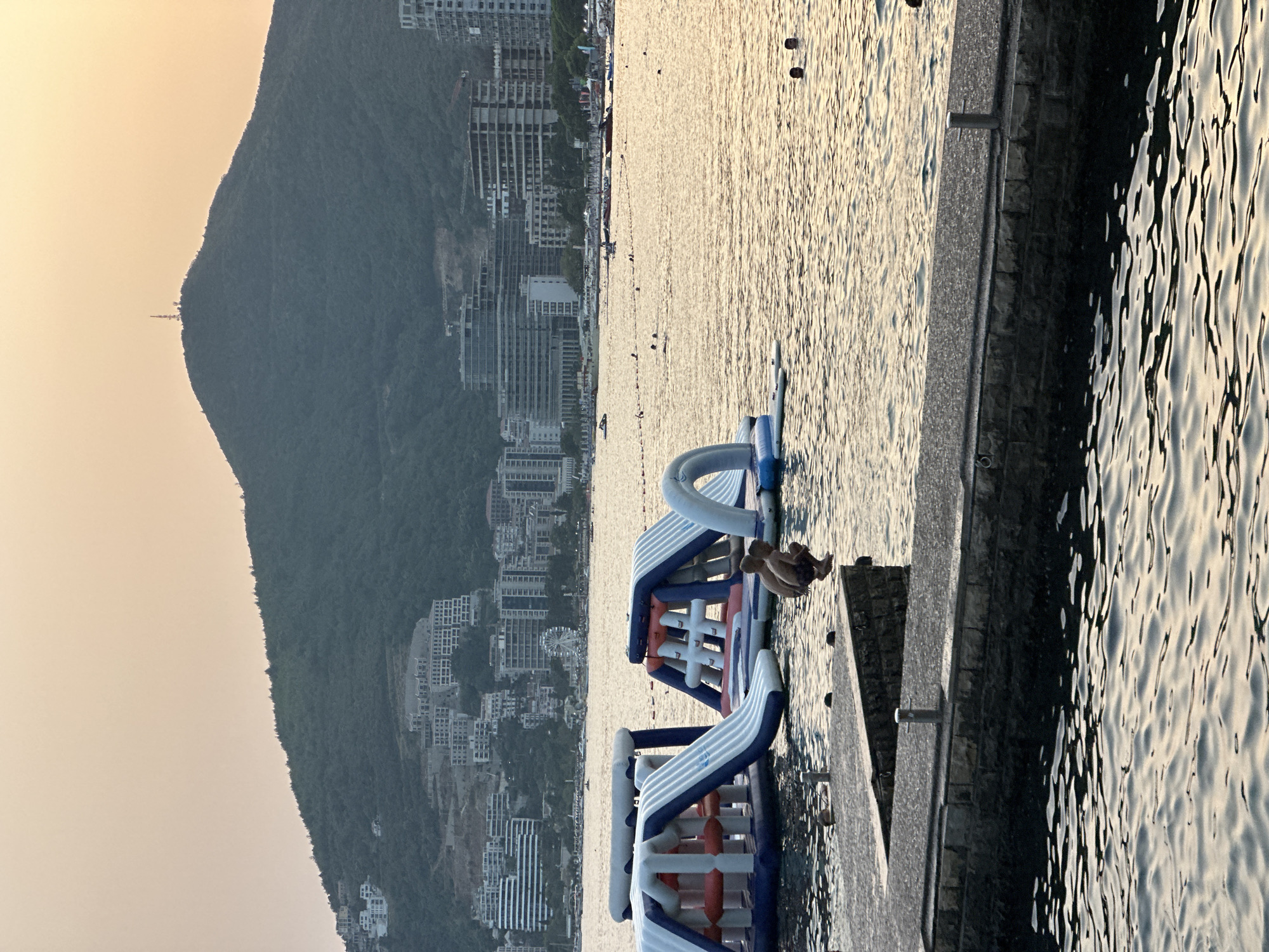 The image captures a scene at Mogren Beach in Budva, Montenegro, during a sunset. In the foreground, a stone pier extends into the calm, shimmering water. On the pier, two individuals, appearing to be young males, are crouched down, seemingly preparing to jump into the sea. One of them is in mid-air, captured in the act of a jump.
Beyond the pier, several large, inflatable water park structures are visible, some featuring slides and climbing elements. The water is populated with other swimmers enjoying the evening. In the midground, the coastline of Budva is lined with numerous hotels and apartment buildings, cascading up the steep, tree-covered hillside. The slopes of the hill are dotted with more buildings and construction sites.
Dominating the background is a large, verdant mountain, its peak slightly obscured by a hazy, pastel-colored sky, indicative of the fading sunlight. A telecommunications tower is perched on the very summit of the mountain. The overall atmosphere is serene, with the golden light of sunset casting a warm glow over the water and the cityscape.