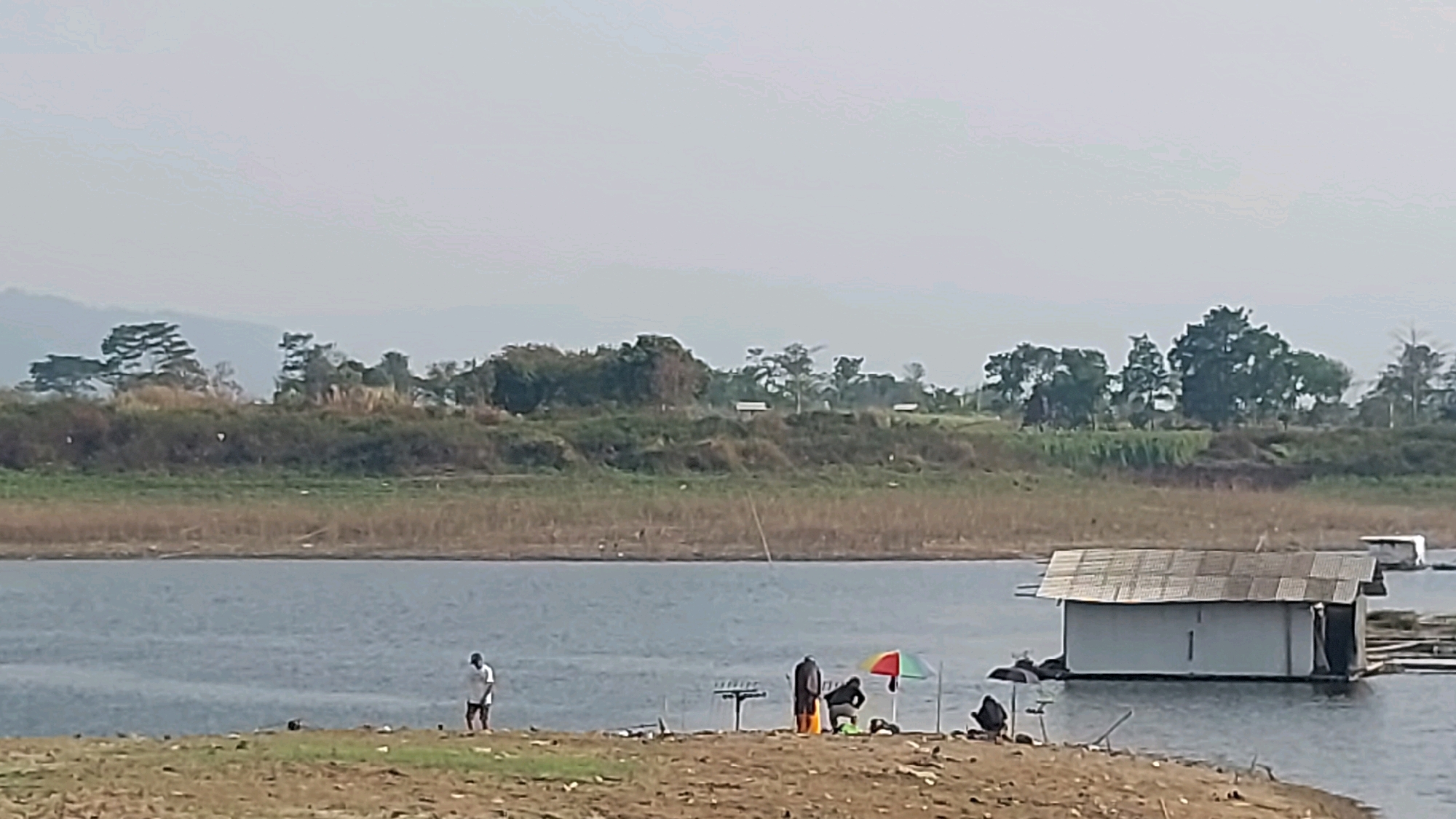 This image depicts a scenic view of a body of water, likely a lake or reservoir, with a shore on the left and a floating structure on the right. The shore features dry, light-brown earth with scattered debris and some green grass. On the shore, there are several individuals and fishing equipment. One person stands alone on the left, appearing to be fishing. Further to the right, a group of three people are gathered. One person is standing, another is seated under a colorful umbrella, and a third is partially visible. Fishing rods are set up on the shore.
In the water, a white, rectangular floating structure resembling a small house or shed with a corrugated metal roof is prominent on the right side of the image. It appears to be moored or anchored. Another similar, smaller floating structure is visible further in the distance to the right.
The background consists of a gentle slope covered with dry grass and vegetation, interspersed with trees. Beyond this, a range of hills or mountains is visible in the distance under a hazy, light-colored sky. The overall atmosphere suggests a calm, possibly rural setting.
The location is identified as Bojonghaleuang, Indonesia, a place known for its natural beauty and potential for water-related activities.