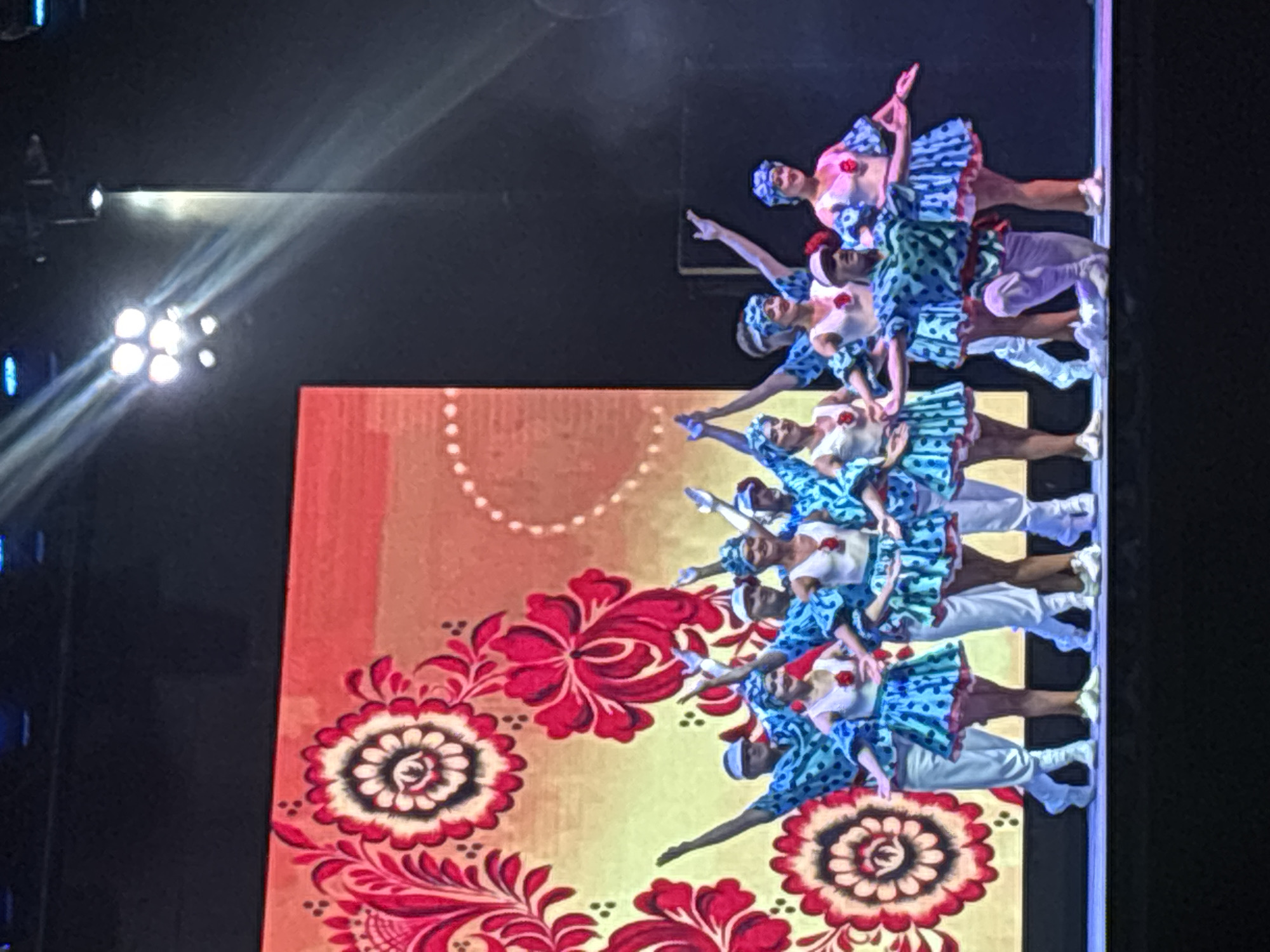 A group of ten performers, dressed in blue and white polka-dot costumes with red accents, are on a stage in front of a large screen displaying a vibrant, floral pattern. The performers are arranged in two rows, with the front row slightly lower than the back row. The front row consists of five individuals, two on the far left and three on the right, with one person kneeling in the center. The back row has five individuals, spaced out behind the front row. All performers are wearing matching headwear and appear to be mid-performance, with some raising their arms in unison. The stage lighting is visible, with spotlights casting beams of light from the upper right corner. The background screen features stylized red and pink floral motifs against a yellow and orange gradient. The overall impression is of a theatrical or cultural performance.