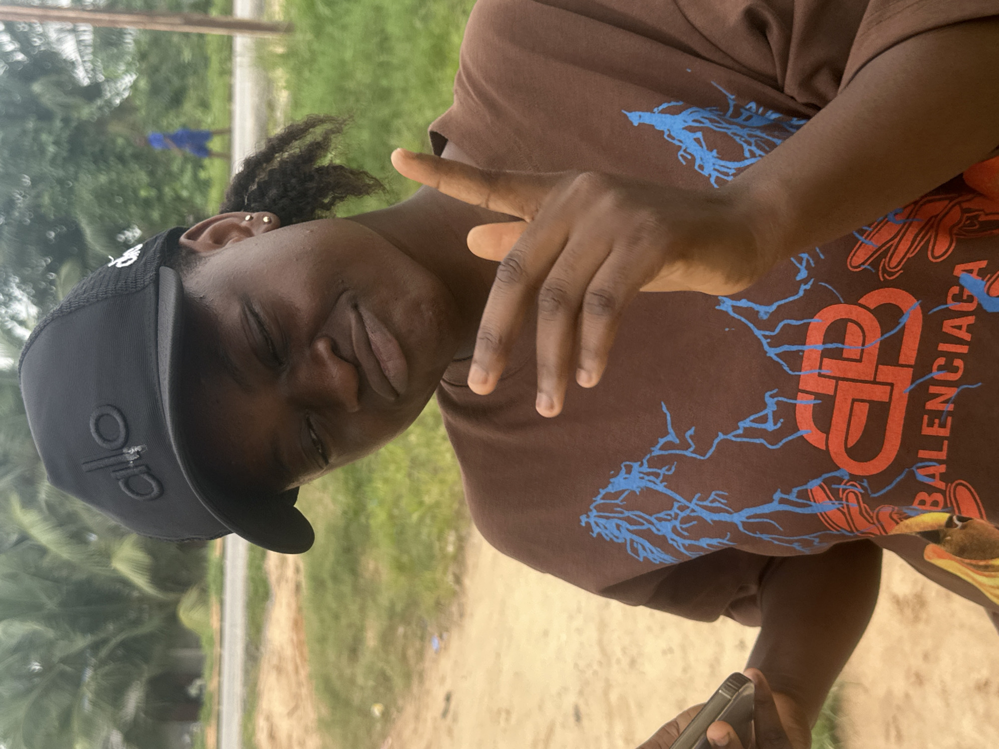 A young Black woman, wearing a black baseball cap with an "alo" logo and a brown t-shirt with a blue lightning bolt design and a red "BB" logo, poses outdoors in Ganyingbo, Nigeria. She is winking and holding up two fingers in a peace sign with her right hand. Her left hand is holding a smartphone. The background consists of greenery, dirt, and a distant figure walking on a road.
