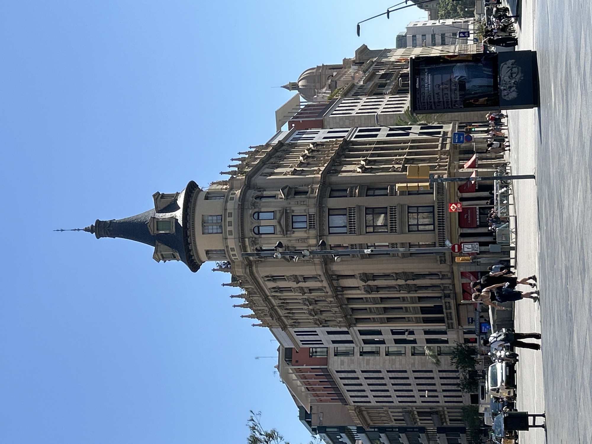 The image shows a street scene in Barcelona, Spain, with a prominent, ornate building in the center featuring a conical tower topped with a spire. The building is cream-colored with decorative elements, multiple windows, and balconies. To the left and right of this central building are other multi-story structures, some with modern facades and others with more classical architecture, including a dome visible further down the street.
The street level shows pedestrians walking on a sidewalk and crossing the street. Several cars and a motorcycle are visible on the road. A large advertising kiosk is positioned prominently in the foreground. The sky is clear and blue, indicating a sunny day. The overall impression is of a busy urban environment in Barcelona, likely a commercial or historical district given the architecture.