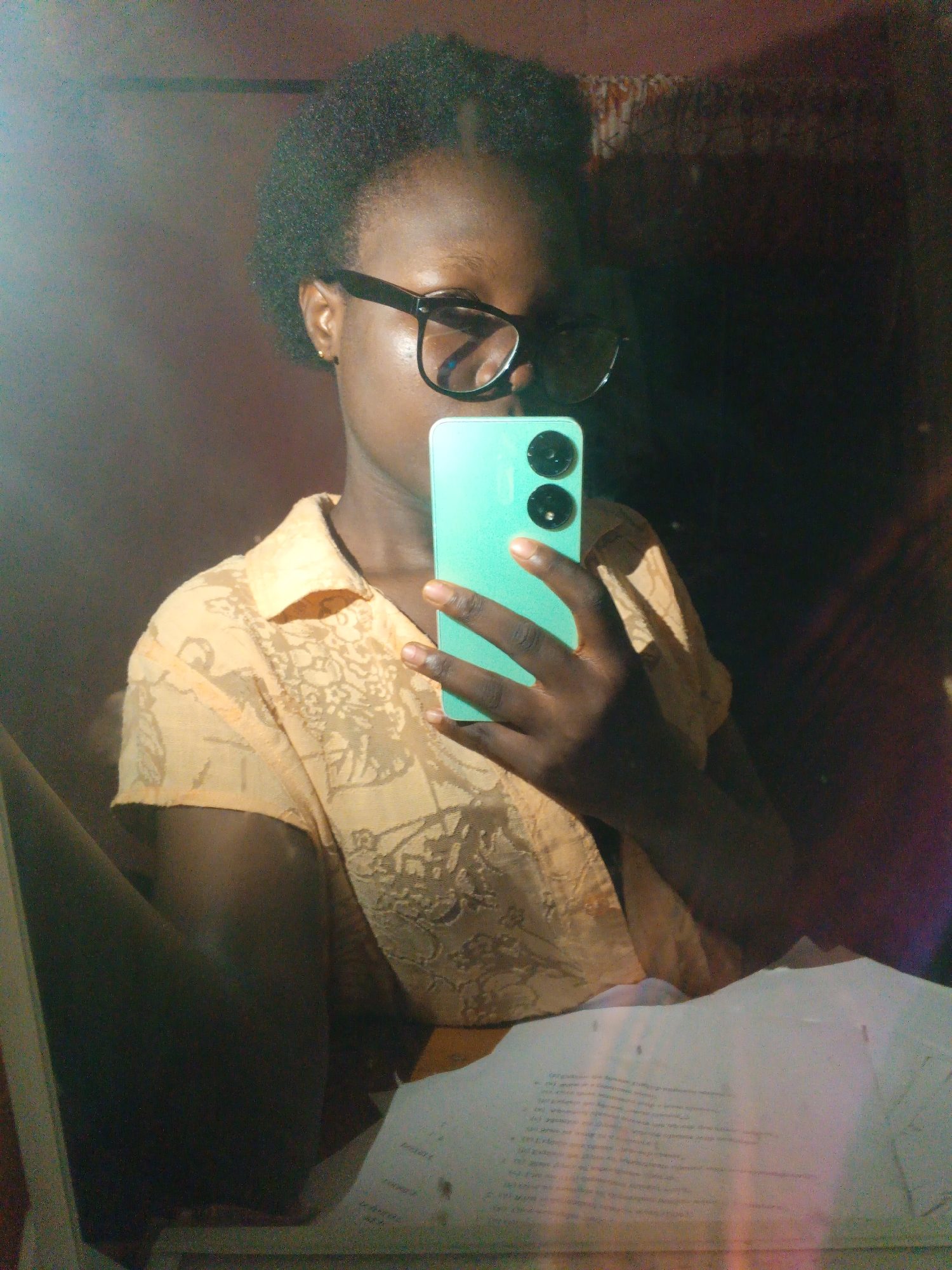 A young Black female wearing glasses and an orange collared shirt is taking a selfie in what appears to be a bedroom in Onitsha, Nigeria. She is holding up a turquoise smartphone with two black camera lenses. The room is dimly lit with some light reflecting off a surface to her left, possibly a mirror or window. The background is dark and indistinct, with hints of patterned fabric or wallpaper. Papers are visible on a surface in the foreground.