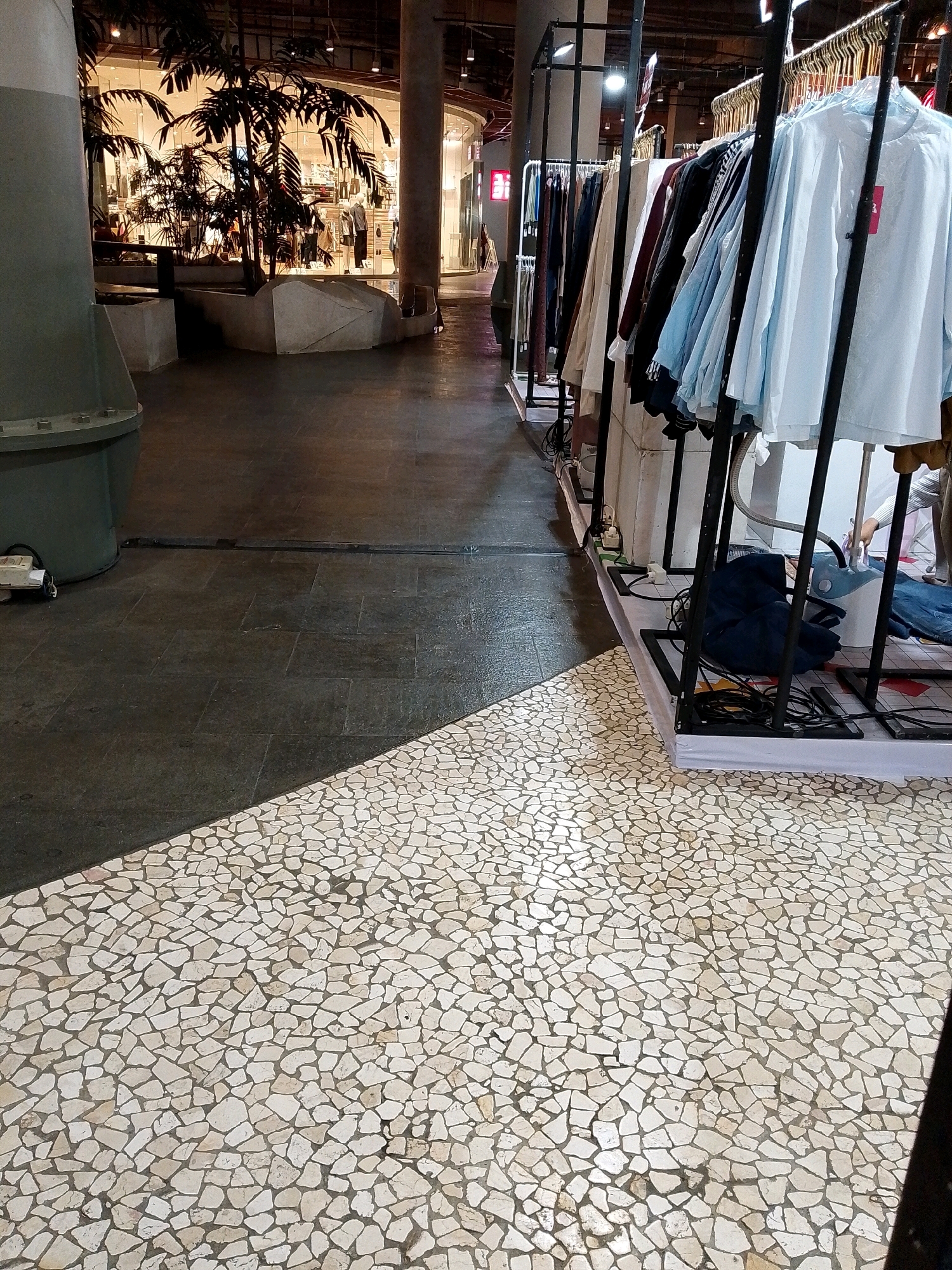 The image depicts an indoor retail environment, likely a clothing store, with a focus on clothing racks filled with garments. In the foreground, the floor transitions from dark, large-tiled flooring to a section of smaller, irregular white and cream-colored stones, creating a distinct visual separation. To the left, a large, dark green cylindrical object is partially visible, possibly a structural element or display. Behind this, a glass-enclosed area with palm plants and mannequins suggests a storefront or interior display. The right side of the image showcases multiple clothing racks with various articles of clothing, predominantly light-colored shirts and dresses, on display. Electrical cords and a garment steamer are visible among the displays, indicating active retail operations. The lighting appears to be artificial, typical of an indoor commercial space. The context of Makassar, Indonesia, is not visually evident within the provided image itself.