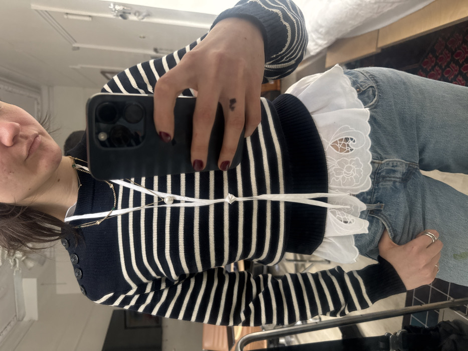 An individual, appearing to be a woman with dark brown hair, is captured in a mirror selfie indoors. She is wearing a navy blue and white horizontally striped long-sleeved sweater with three dark buttons on the left shoulder. Beneath the sweater, a white shirt with an embroidered eyelet lace scalloped hem is visible. She also wears light blue denim jeans, with her left hand placed in a pocket, displaying two silver-toned rings. Multiple gold-toned necklaces and a long white lanyard-style necklace with a central knot and decorative ends are worn around her neck. Her right hand, featuring dark red nail polish and a small dark tattoo on the inner middle finger, holds a dark-colored smartphone (likely an iPhone) to take the photograph. The reflected background reveals an interior residential space consistent with an apartment in New York City, United States of America, featuring a light-colored wall, a dark table or desk with a chair, a framed object on the wall, and a glimpse of a window with white textiles to the right. The ceiling exhibits crown molding.