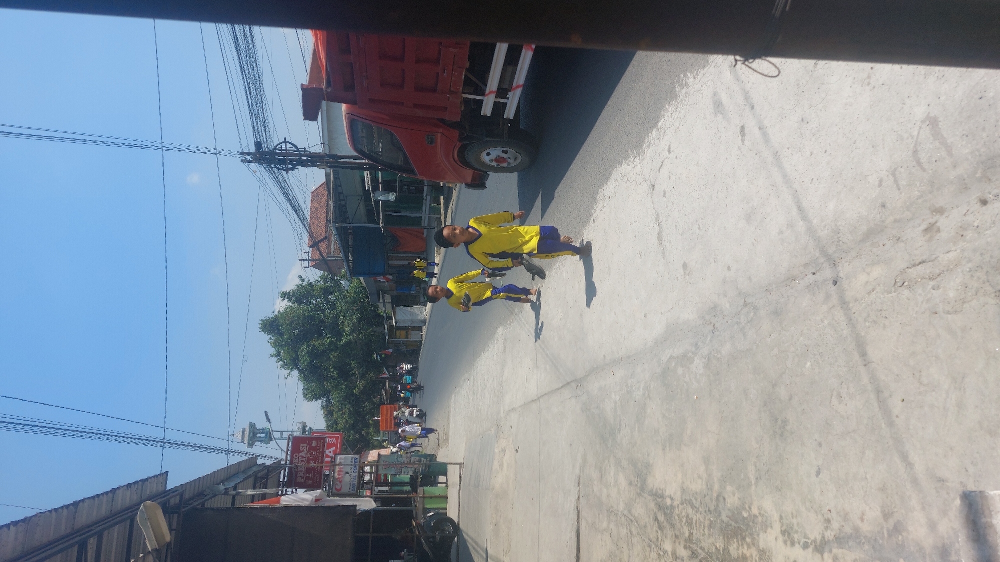 The image captures a sunny day on a street in Karas, Indonesia. Two young boys, dressed in matching yellow long-sleeved shirts and blue pants, are running on the sidewalk. The boy in the foreground is slightly ahead of the one behind him. They are both barefoot and appear to be carrying their shoes. To the right of the boys, a red truck is parked on the road. The street is lined with buildings on the left, some with awnings and signage, and more buildings and trees are visible in the background. Electrical wires are strung across the sky. The scene suggests a typical day in a town with residential and commercial activity.