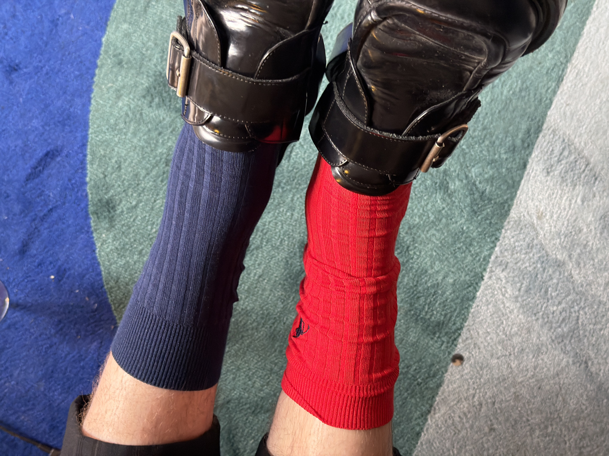 A top-down view shows a person's lower legs and feet, shod in black patent leather shoes with buckled straps. The person wears a navy blue ribbed sock on the left foot and a red ribbed sock on the right foot. The background is a striped carpet in shades of blue, green, and white. There is no evidence to identify the location as London, United Kingdom.