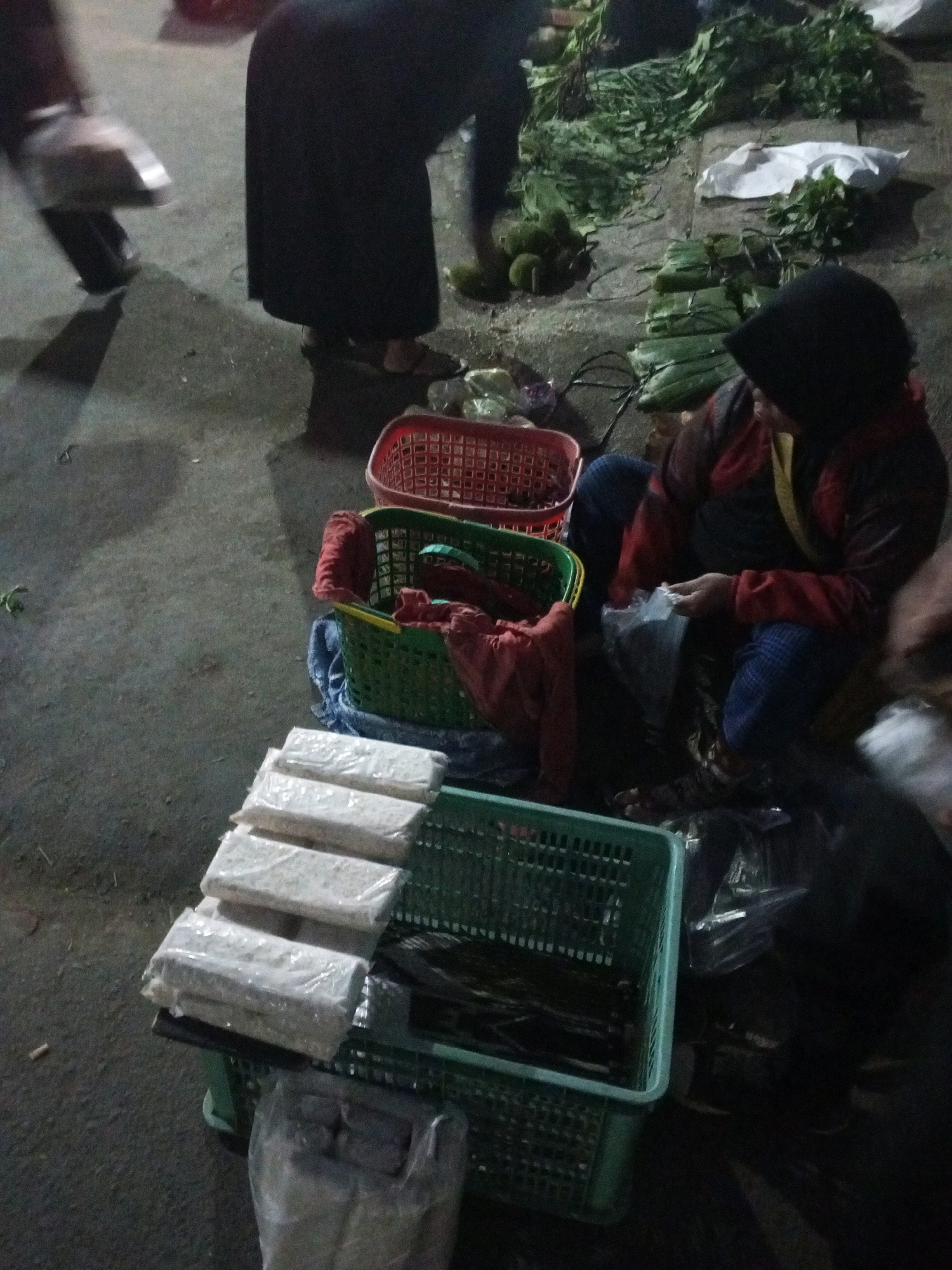 This image captures a street market scene in Gentan, Indonesia, with a focus on food products and the vendors. In the foreground, several rectangular packages, likely tempeh, are neatly stacked and wrapped in transparent plastic, resting in a green plastic basket. To the right of these packages, a person wearing a dark head covering and a red and black jacket is visible, appearing to be engaged in a transaction or preparing items. Their hands are holding a plastic bag.
Behind the packaged food, other market elements are discernible. A green basket contains a red cloth. Further back, a red basket sits on the ground. The ground itself appears to be paved or a compacted surface typical of an outdoor market. In the background, a pile of fresh green vegetables, possibly leafy greens and other produce, is spread out, indicating a variety of goods available. A person in dark clothing with a distinct metallic object on their left foot, possibly a shoe or footwear, is partially visible on the left side of the image, suggesting movement or a vendor’s presence. The lighting suggests it might be evening or an overcast day.