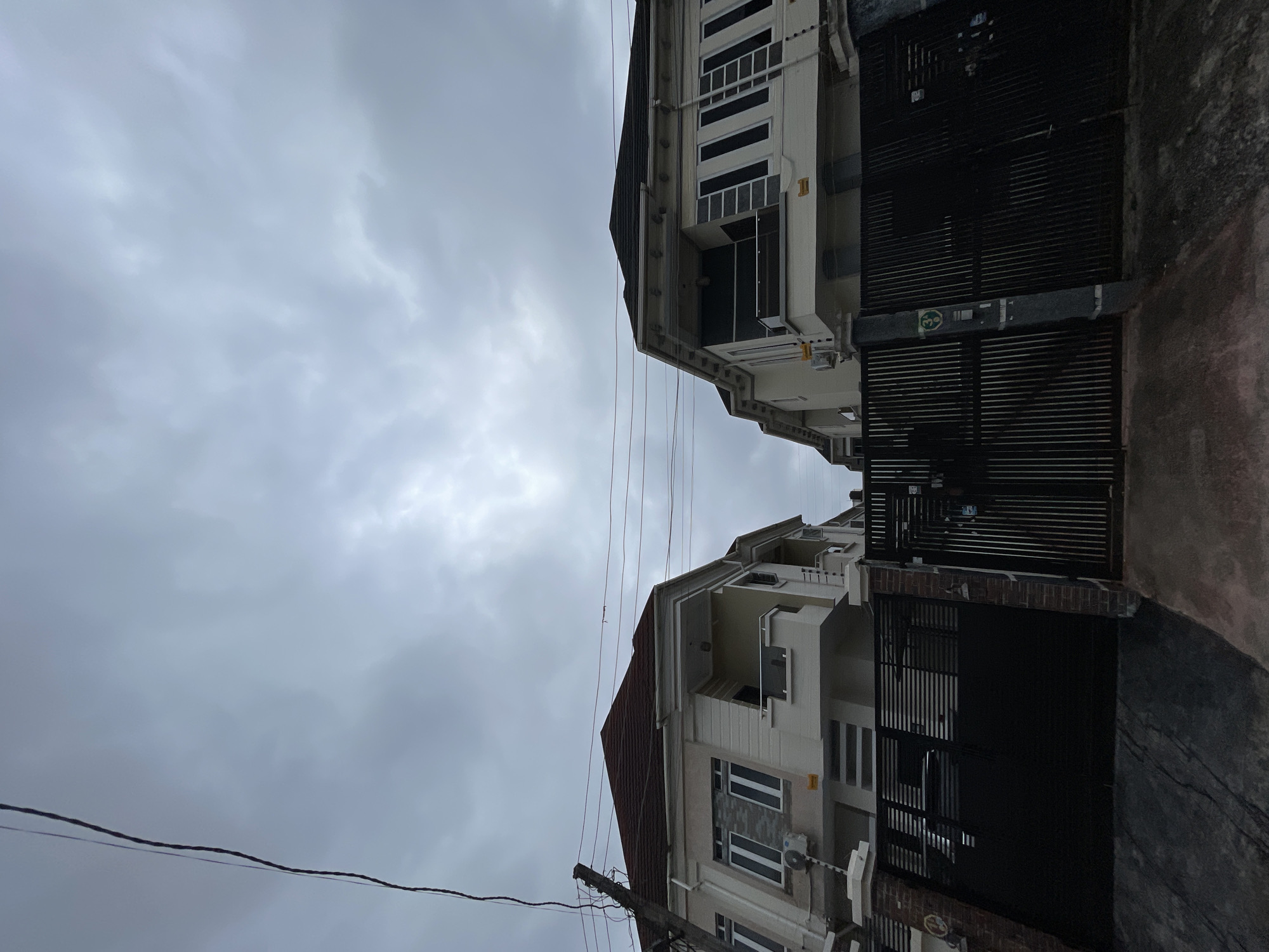 The image shows a cloudy sky over two modern houses with black metal gates in front of them, likely in Ado, Nigeria. The houses appear to be two-story structures with beige exteriors, white trim, and dark-colored roofs. Both have balconies and multiple windows. The gates are made of horizontal and vertical metal bars. A utility pole with wires is visible on the left side of the image. The ground in front of the gates appears to be paved.