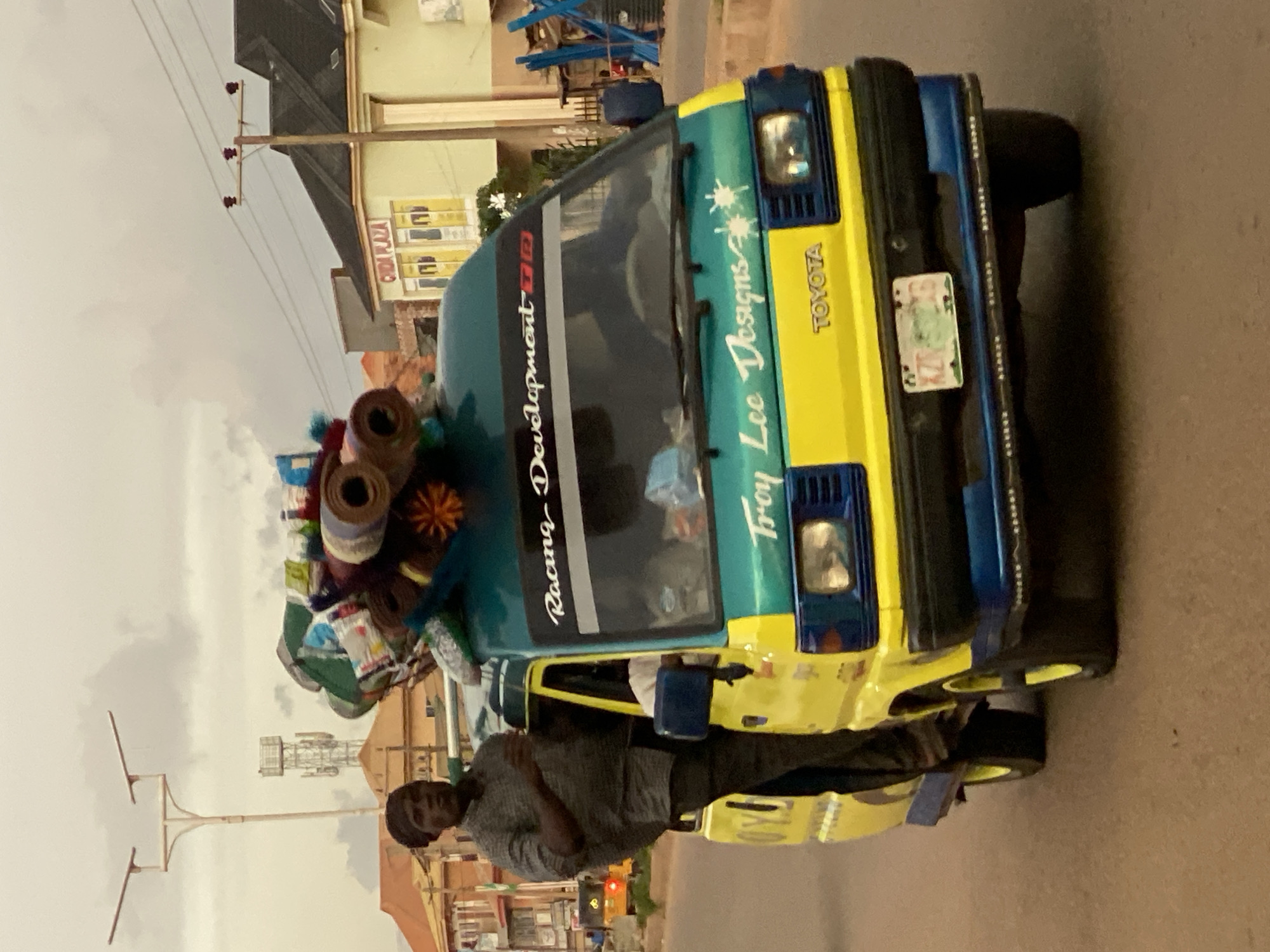 In Sabon Tasha, Nigeria, a yellow and teal Toyota Hiace van is traveling on a paved road. The van is loaded with various goods on its roof, including rolled-up carpets or mats and other bundled items. A man stands in the open doorway of the van, appearing to hold onto the roof. The van's front features "Racing Development" written on the windshield and "Troy Lee Designs" with decorative starbursts on the teal section above the headlights. A "TOYOTA" emblem is visible on the yellow portion. The van has a license plate with the characters "KZR 506 DG". In the background, buildings, streetlights, and utility poles are visible. The sky is overcast.