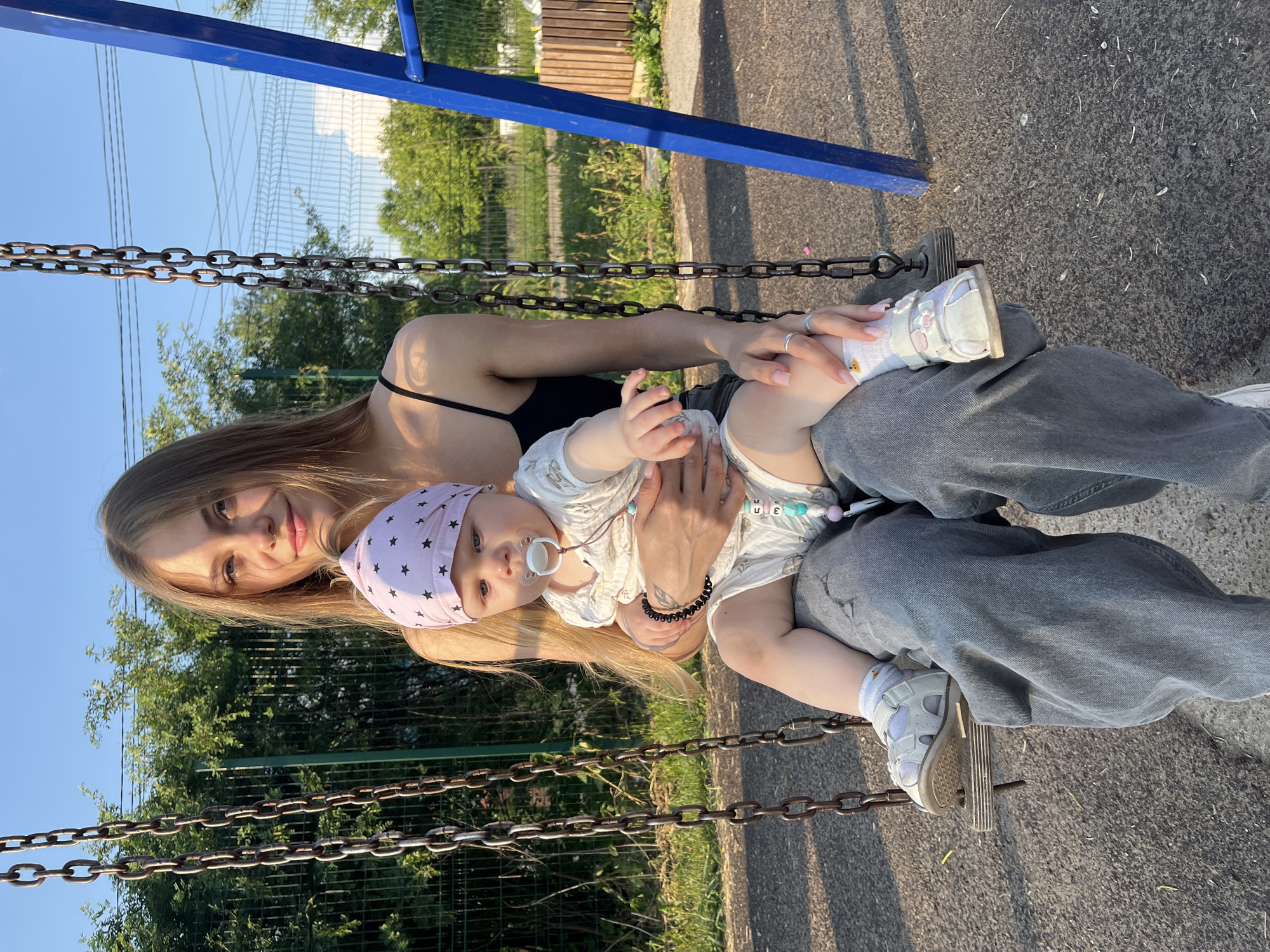 A young woman with long blonde hair sits on a swing, holding a baby in her lap. The woman wears a black tank top and grey jeans. The baby is dressed in a white outfit and a pink bandana with stars. The baby has a pacifier in its mouth. They are on a playground with a blue swing set frame. The background shows green trees and a fence. The ground is paved with asphalt. The lighting suggests it is late afternoon or early evening. The image was taken in the city of Fokino, Russia.