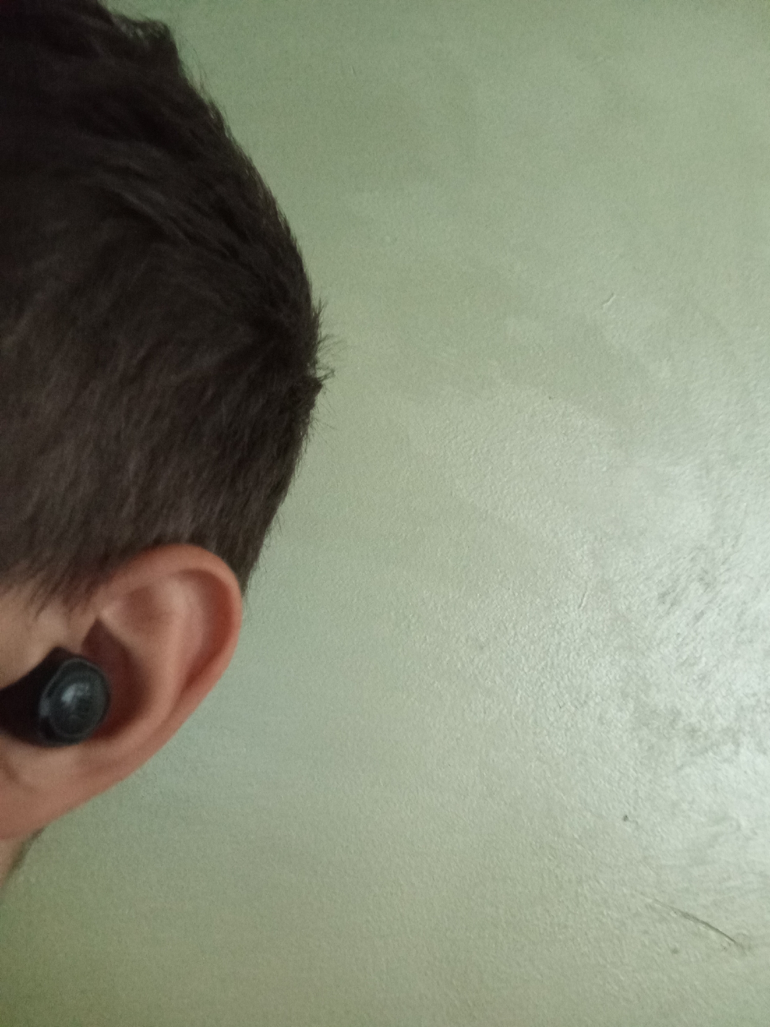 The image shows a close-up view of a person's ear with a black earbud inserted. The person's dark brown hair is visible in the upper left portion of the frame. The background is a textured, pale green wall. The image does not contain any elements that would allow for the determination of its location in Malko Chochoveni, Bulgaria, or any other specific geographical area. The lighting appears to be indoor and diffused.
