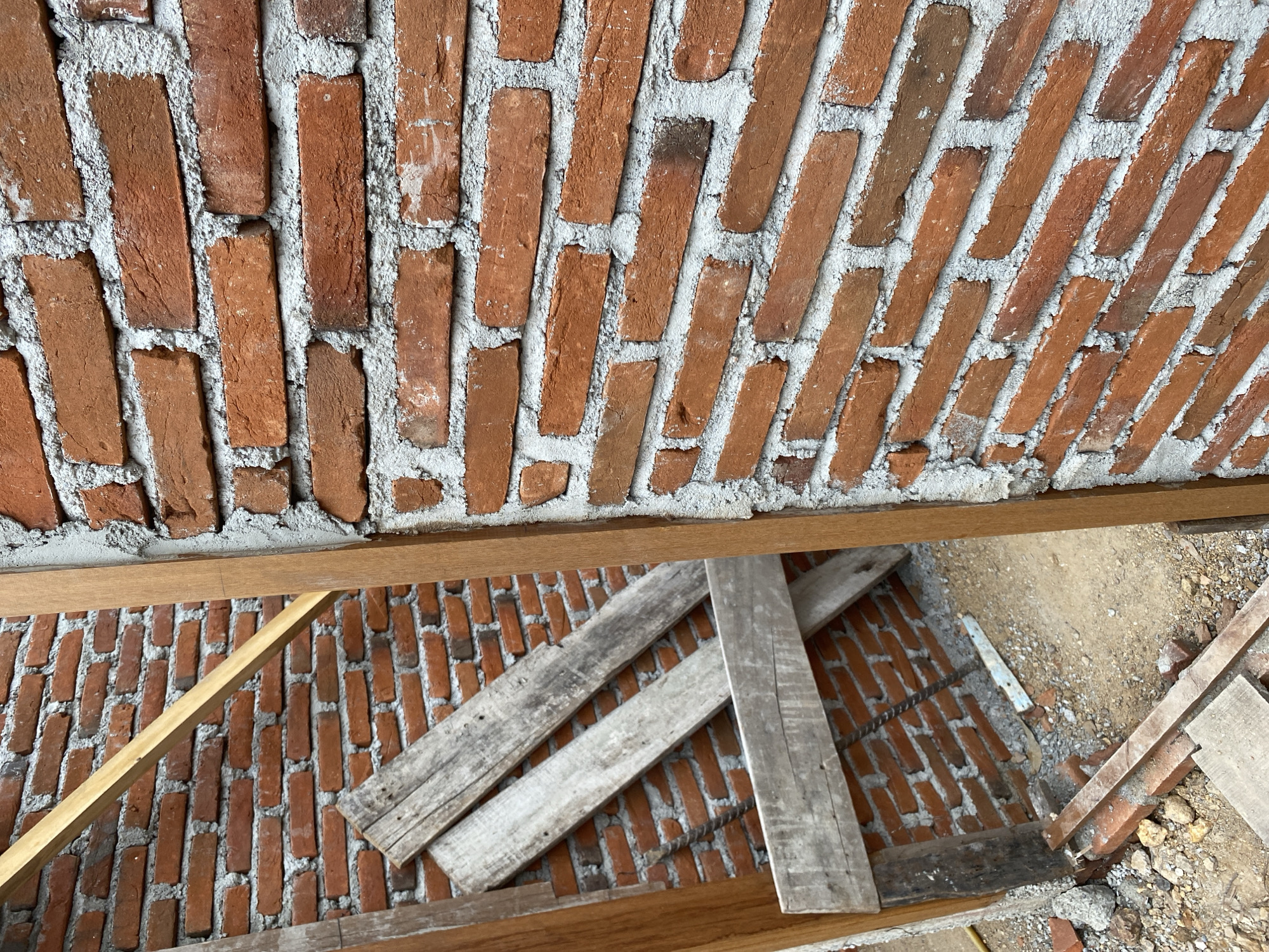 This is a close-up, eye-level, outdoor, full shot of an unfinished brick wall construction in Banda Aceh, Indonesia. The wall is made of red bricks mortared together with gray cement. Wooden planks are propped against the wall, suggesting ongoing construction or repair work. The ground beneath the wall is covered with dirt, small rocks, and scattered brick fragments. The image captures the raw texture and detail of the building materials. There are no people visible in the frame.