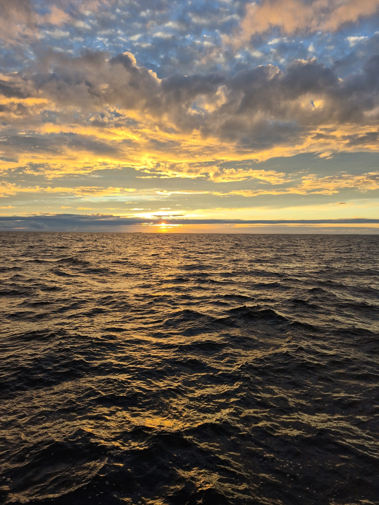 A wide-angle, eye-level shot captures a serene ocean scene at sunset. The sky is a canvas of vibrant colors, with streaks of yellow, orange, and blue clouds illuminated by the setting sun. The sun itself is a small, bright orb just above the horizon, casting a golden glow across the water. The ocean surface is a dark, rippling expanse, reflecting the warm hues of the sky. The waves are gentle, creating a sense of peace and tranquility. There are no people or man-made objects visible in the image, emphasizing the natural beauty of the seascape. The overall impression is one of calm and quiet contemplation, as the day draws to a close.