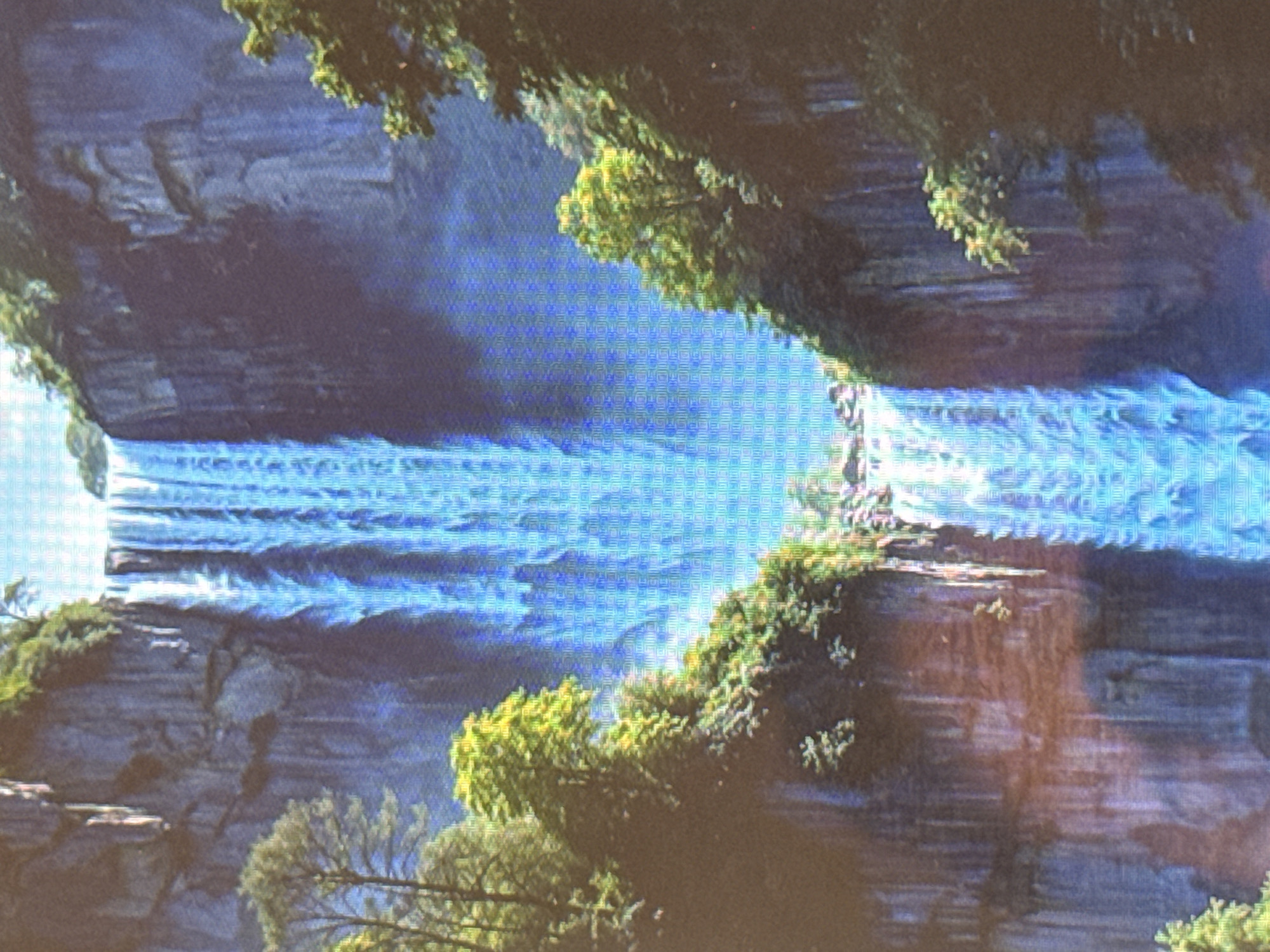 The image displays a majestic waterfall cascading down a sheer cliff face. Lush green trees and foliage frame the sides of the waterfall, with rocky outcrops visible on the cliff. The water itself appears bright blue, with mist rising from the base. The scene is lit by natural light, suggesting an outdoor environment. The image quality is somewhat pixelated.