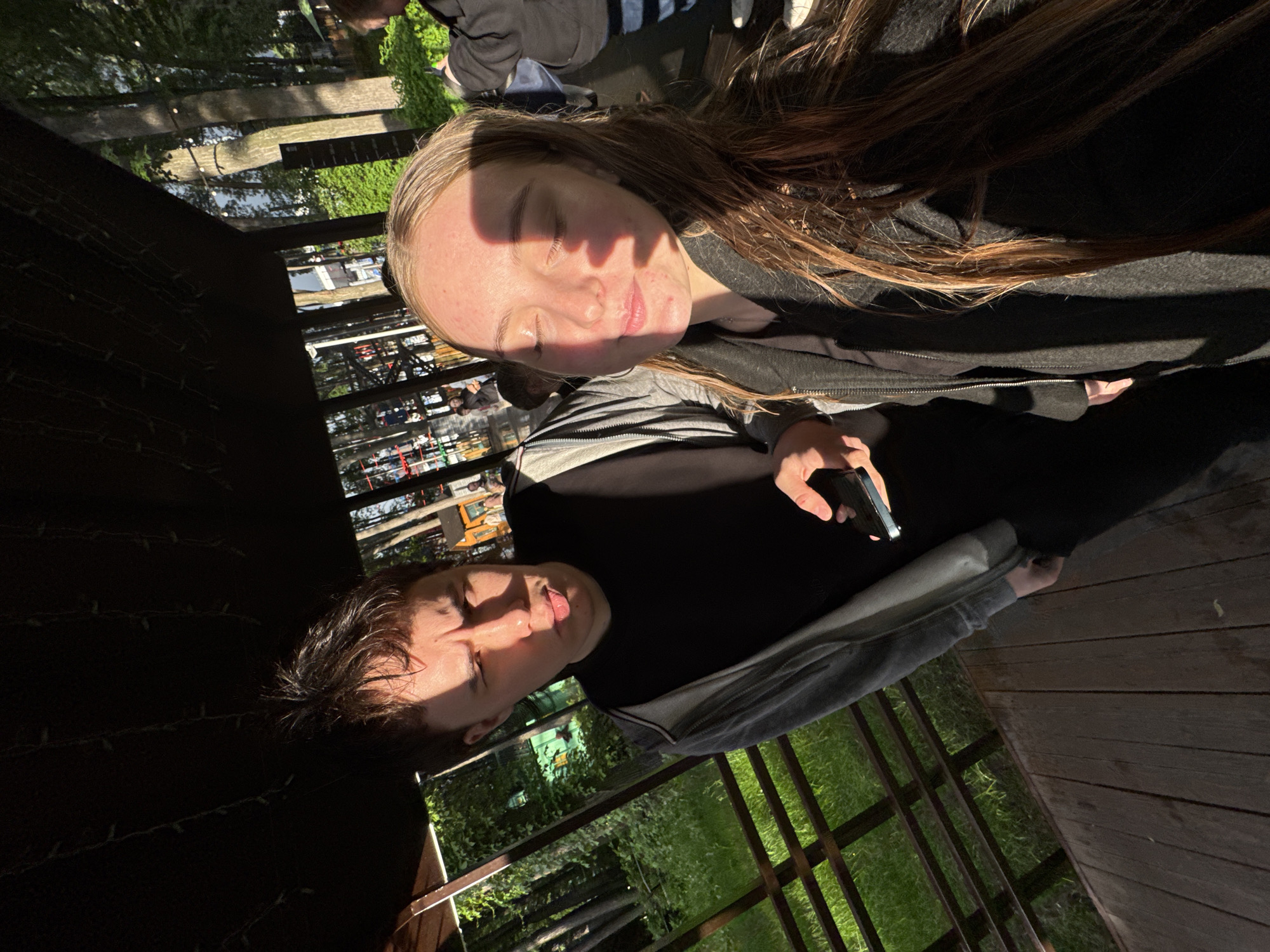 Two people are in a shadowed, outdoor area. The person on the left, with short dark hair and a black shirt, is on a wooden platform. They have a grey jacket. The person on the right has long hair. Both faces are obscured by heavy shadows. The location is possibly a raised wooden platform with metal railings overlooking foliage. The location in Perm, Russia, cannot be determined from the image.