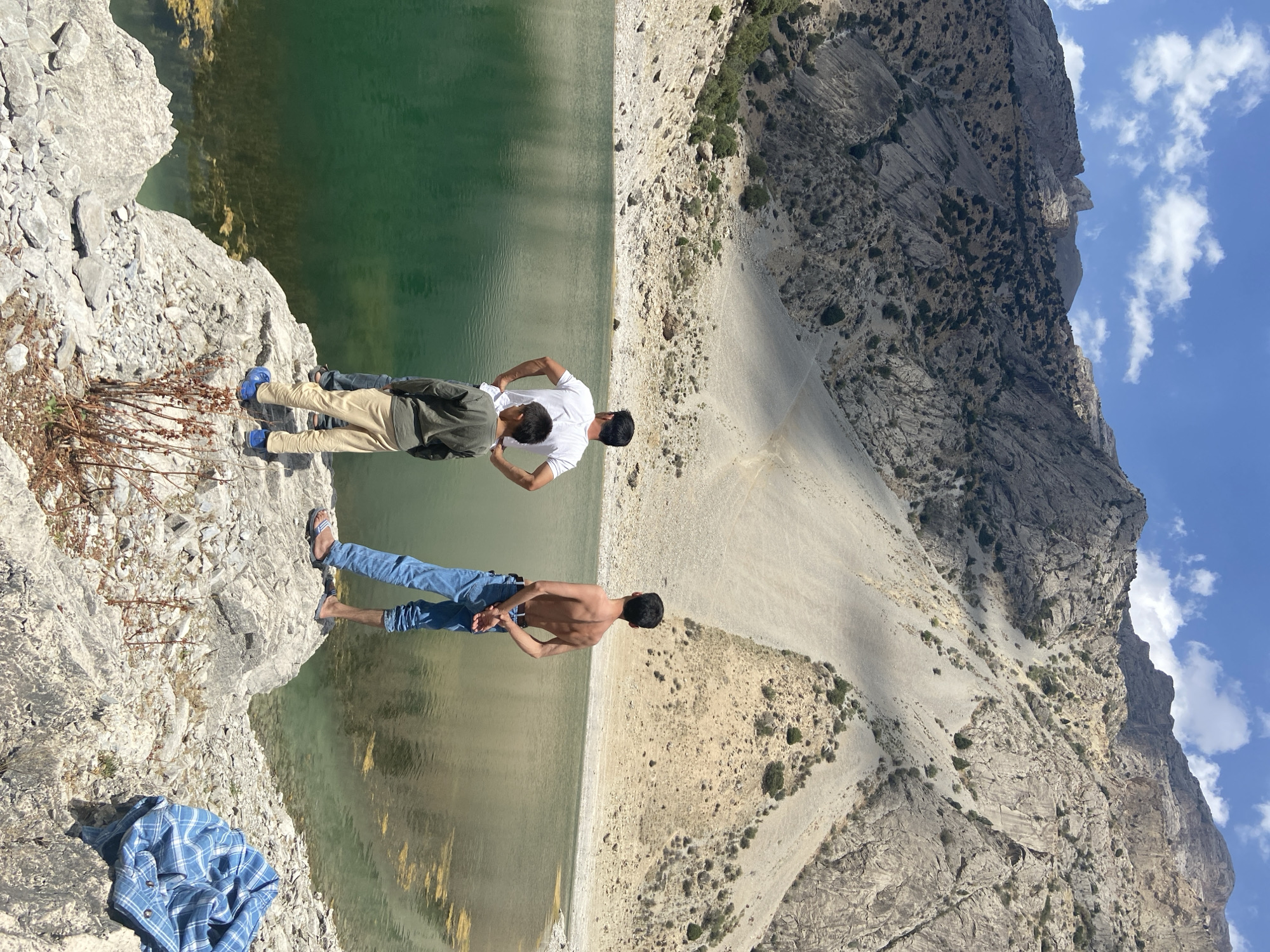 This image captures a serene moment at the Hazratchi Mountains in Ghazza, Tajikistan. Three individuals, two adults and a child, stand on a rocky outcrop overlooking a tranquil, emerald-green lake. The child, wearing a long-sleeved olive shirt and beige pants, stands to the left, his hands behind his back. Beside him, an adult in a white t-shirt and dark pants also has his hands behind his back, his posture relaxed. To the right, another adult, shirtless and wearing blue jeans and flip-flops, faces away from the camera, his hands clasped behind his back. The background is dominated by majestic mountains, their slopes a mosaic of rocky terrain and sparse vegetation, with a bright blue sky dotted with fluffy white clouds overhead. The lake's surface reflects the surrounding landscape and the sky, creating a sense of depth and tranquility. A blue and white checkered shirt lies discarded on a rock to the right of the frame, adding a casual, lived-in feel to the scene. The overall impression is one of peaceful contemplation amidst the grandeur of nature.