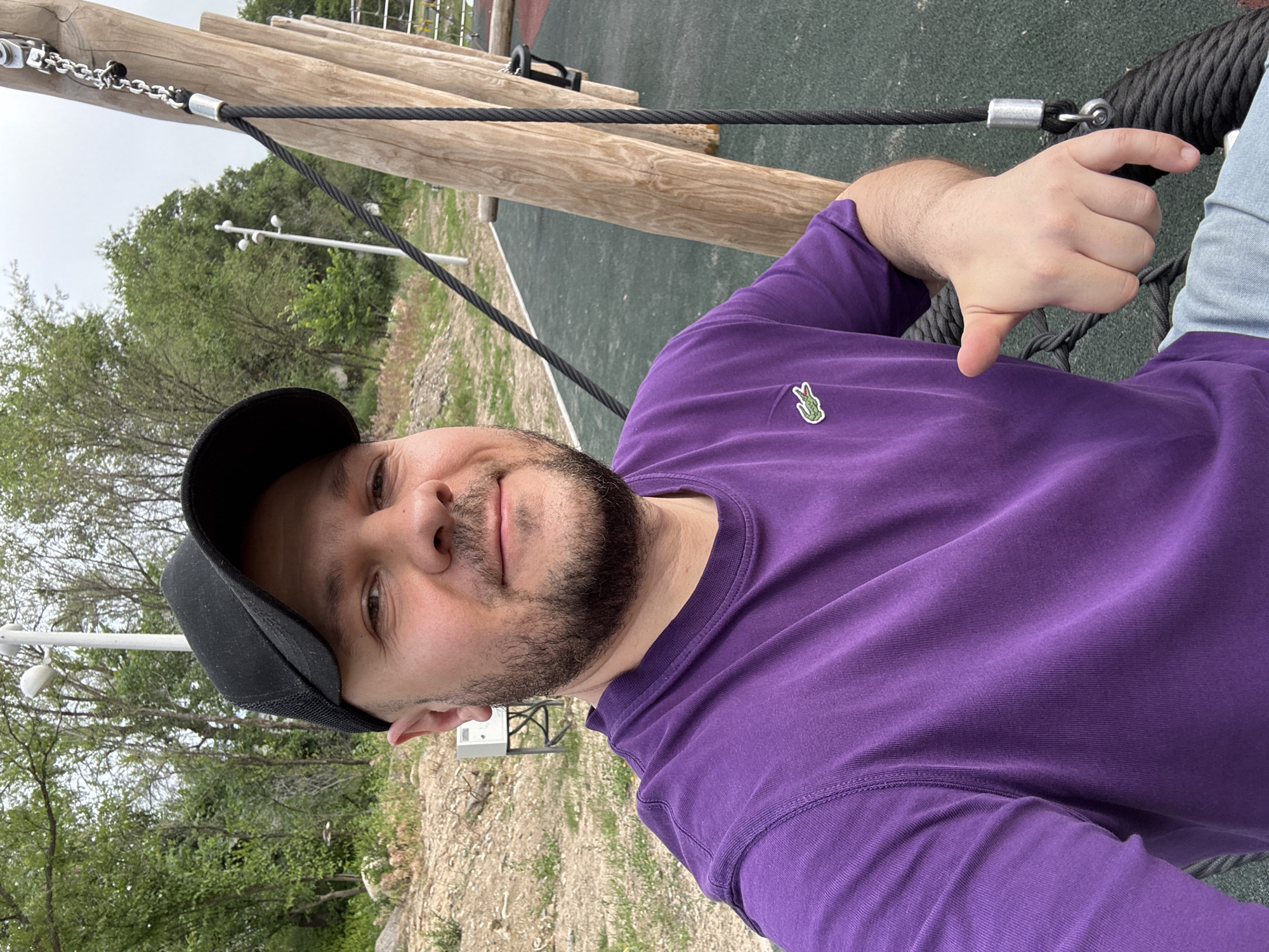 A man wearing a black baseball cap and a purple Lacoste t-shirt is taking a selfie on a swing. He has a beard and is giving a thumbs-up to the camera. The swing is part of an outdoor playground, likely in a park or recreational area. In the background, there are trees, a light pole, and playground equipment. The ground appears to be covered with a rubberized surface. The scene is set against a slightly overcast sky. This image was taken at the Kok Tobe park in Almaty, Kazakhstan.