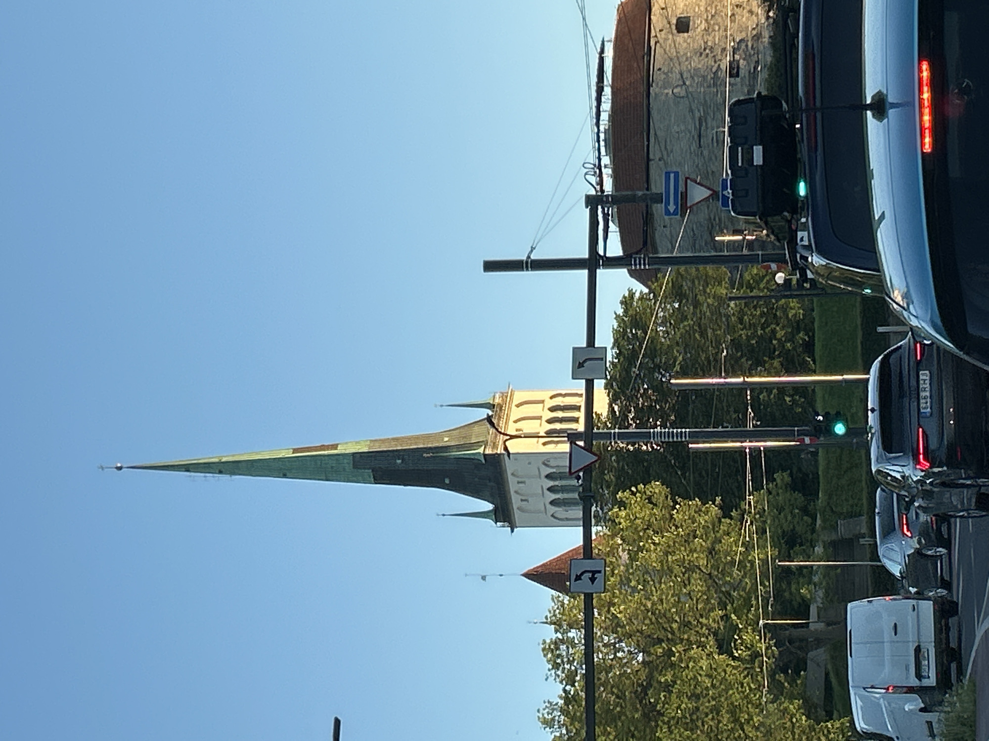This image captures a street scene in Tallinn, Estonia, featuring the prominent spire of St. Olaf's Church (Oleviste kirik) in the background under a clear blue sky. The church's tall spire is green, tapering to a point topped with a cross and an orb, and its cream-colored tower exhibits multiple arched windows. To the left of the church, a smaller building with a red conical roof is partially visible, while to the right, a round, stone-built tower with a reddish-brown roof, consistent with the city's ancient fortifications, is present. Dense green trees are interspersed among the buildings and foreground structures.
In the foreground, a road occupied by multiple vehicles is visible. On the far left, the rear of a white van (Ford Transit-style) is stationary. Centrally, a dark sedan, bearing the license plate "546 RHJ", is also visible from the rear. Dominating the immediate foreground on the right is the rear of a light blue/teal SUV, which has a dark, rectangular cargo carrier mounted on its roof rack; its central red brake light is illuminated. A green traffic light is active on the right side of the road. Utility poles and street light poles are interspersed, supporting horizontal bars from which multiple traffic signs hang, including a black square sign with a white left-turn arrow, an inverted red-bordered triangle "Give Way" sign, and a blue rectangular sign with a white left-pointing arrow. The scene is brightly illuminated by natural sunlight, suggesting either morning or late afternoon, with shadows cast to the left.