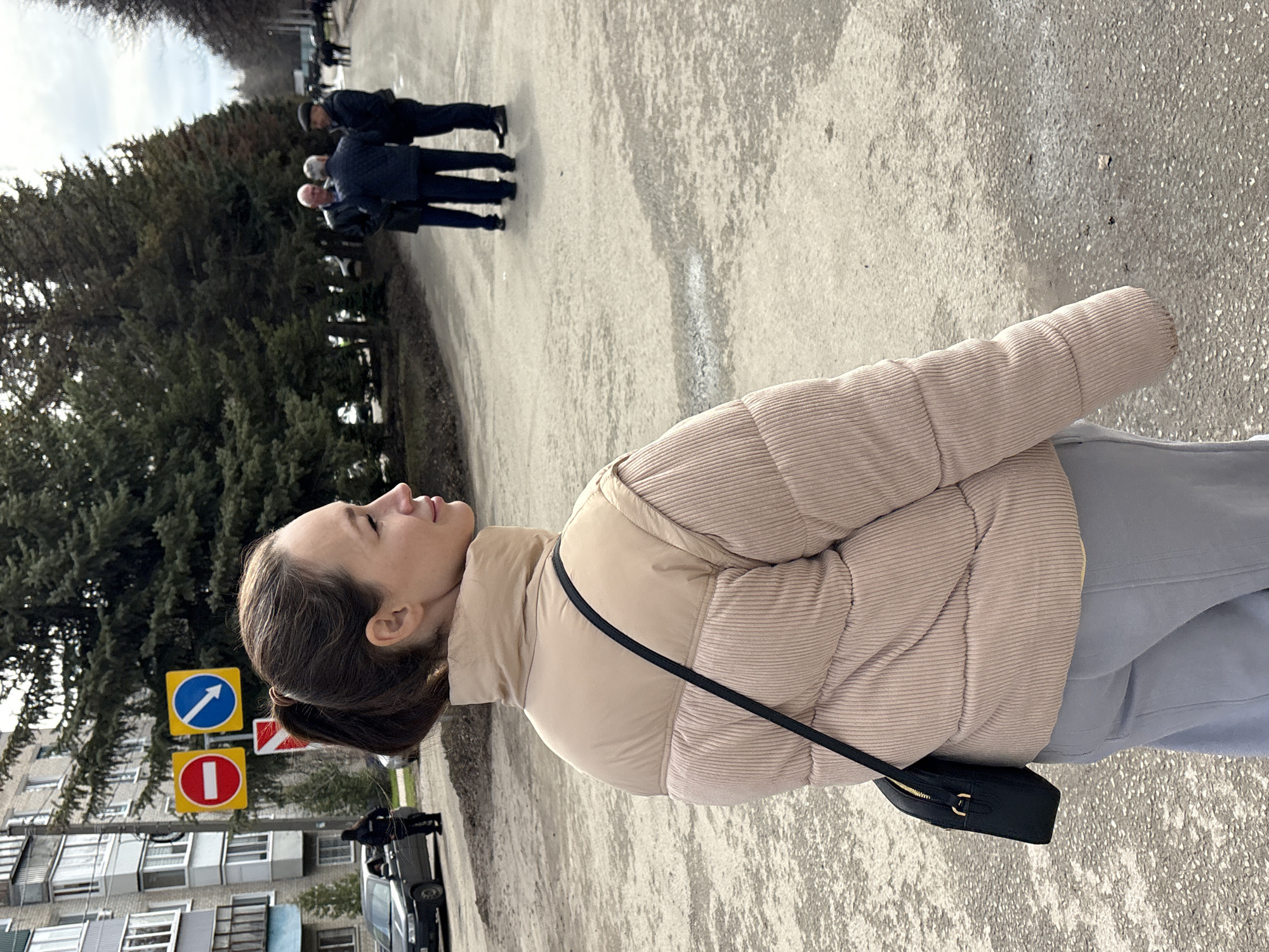 The image depicts a woman standing outdoors. She has brown hair pulled back into a ponytail, and wears a beige puffer jacket and light blue pants. A black purse with a strap is slung across her body.
In the background, there is a street with a building on the left and trees to the left and center. Traffic signs are visible near the building: one blue with a white arrow pointing upwards and to the right, and one red with a white horizontal bar. A black car is parked nearby. Further away, a group of people are seen walking on the sidewalk.
The location is Ulyanovsk, Russia, based on the road signage in the image.