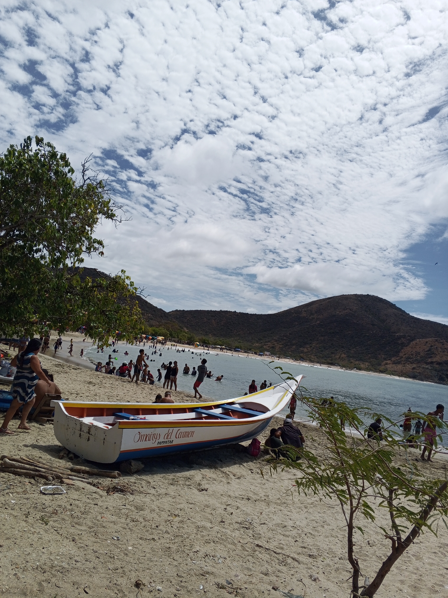 The image captures a vibrant beach scene. In the foreground, a white boat with yellow and blue trim rests on the sandy shore, its side displaying "Omaury's del Carmen PAMPATAR." To the left of the boat, a few logs are scattered, while to the right, a young, leafy tree grows.
The midground is crowded with people enjoying the beach and water. Some are swimming, wading, and others are sitting or standing along the shoreline. The beach slopes gently towards the water, and there are other boats and objects partially visible.
In the background, rolling hills rise up from the edge of the beach, covered with vegetation. The sky above is filled with a mix of white clouds and patches of blue sky, contributing to a bright and sunny atmosphere.
The exact location is not specified, only PAMPATAR is printed on the boat, though no city is mentioned.