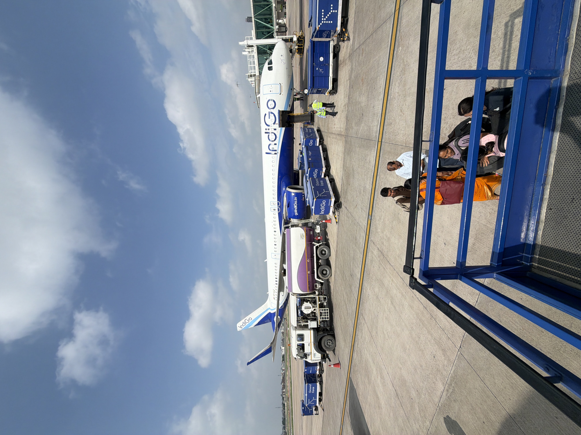 An Indigo Airlines airplane is parked at an airport tarmac in Vikram Nagar, India, under a partly cloudy sky. The plane, with the airline's blue and white livery, is connected to a jet bridge on the right side of the frame. In front of the aircraft, a white fuel truck with purple accents is parked next to several blue baggage carts, some of which are labeled "Indigo" and "Ground Force." Two airport workers in high-visibility vests are near the baggage carts. In the foreground, passengers, including women in traditional Indian attire and carrying luggage, are visible near a blue ramp structure. The ground is paved concrete, with yellow and white markings.