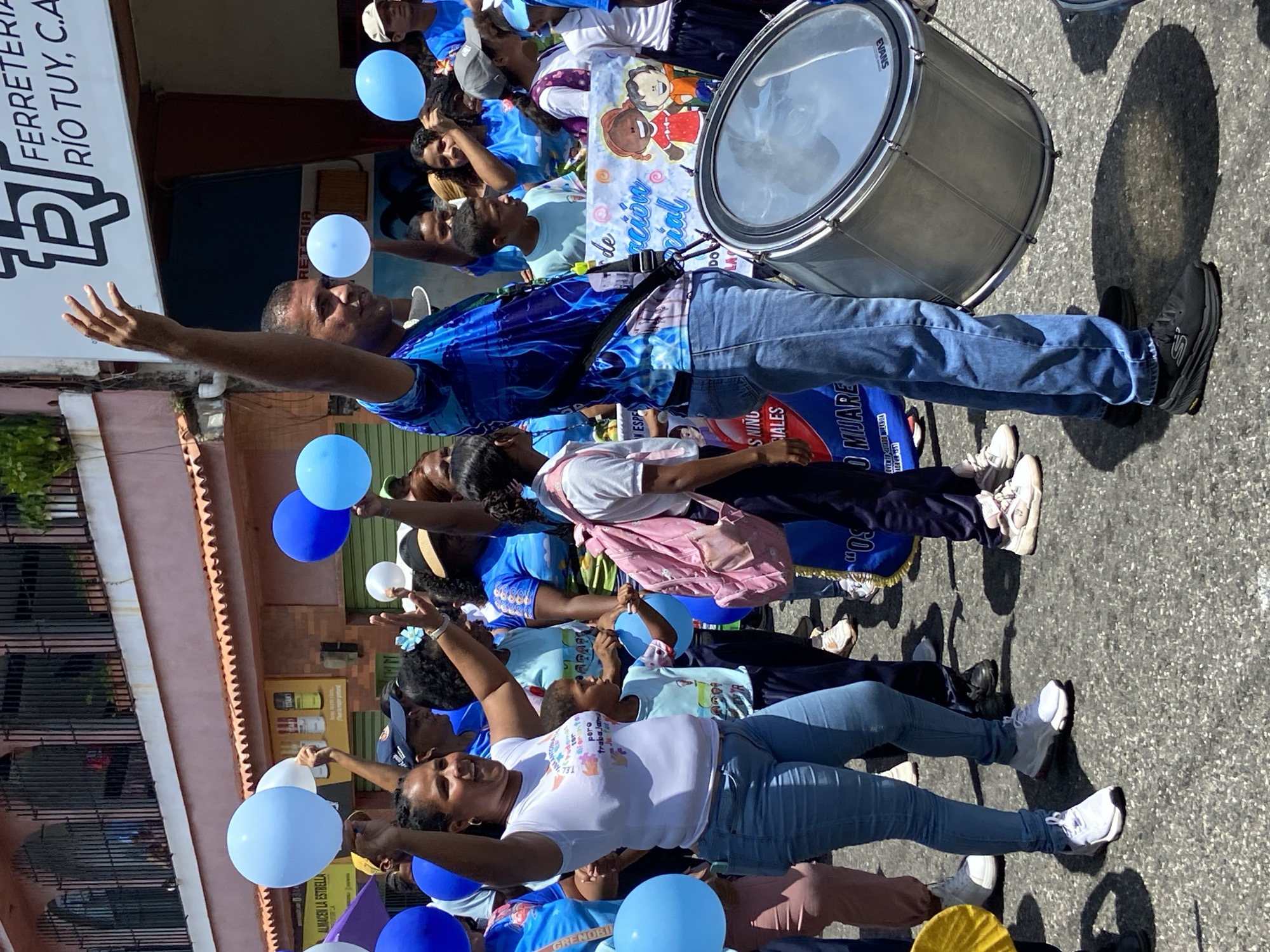 A street-level photograph captures a parade scene on a sunny day in San José de Barlovento, Venezuela. The crowd, which consists of men, women, and children, are dressed in matching blue clothing, with some also wearing white. They wave blue and white balloons, and appear to be marching.
In the background, a silver drum can be seen. A banner with cartoon images and Spanish text is visible as well. Storefronts line the street, displaying signs in Spanish. The street itself is paved and shaded.