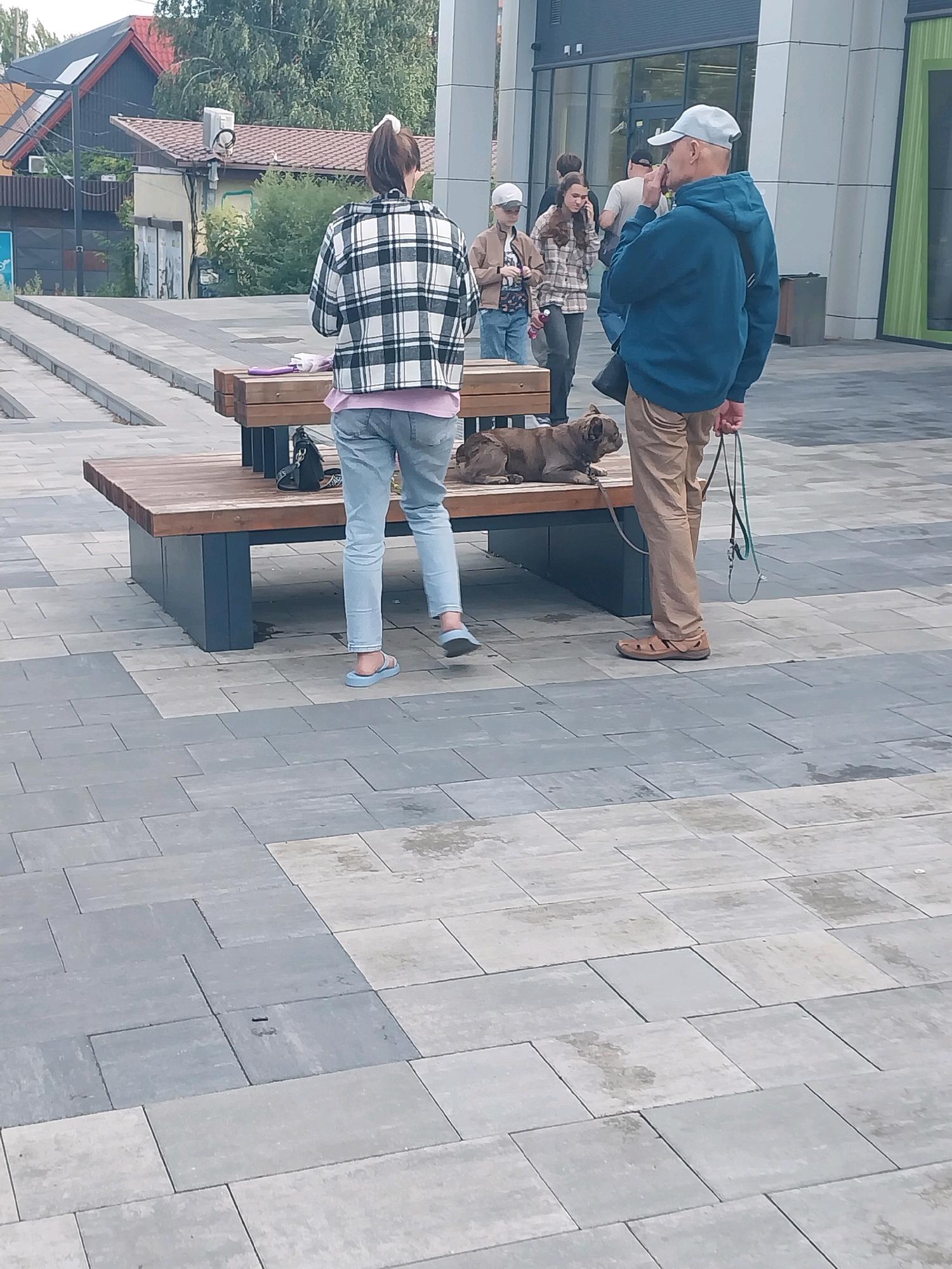 The image depicts a man and a dog in front of a modern building. The man, wearing a blue hooded sweatshirt and tan pants, stands on a paved area, holding a leash attached to a brown French Bulldog lying on a wooden bench. Behind him, a woman in a black and white plaid jacket and light blue jeans stands with her back to the camera. Several other individuals are visible in the background. The environment appears to be an urban outdoor space, possibly a plaza or pedestrian area, with a focus on contemporary architecture and paving. The scene is captured in daylight.