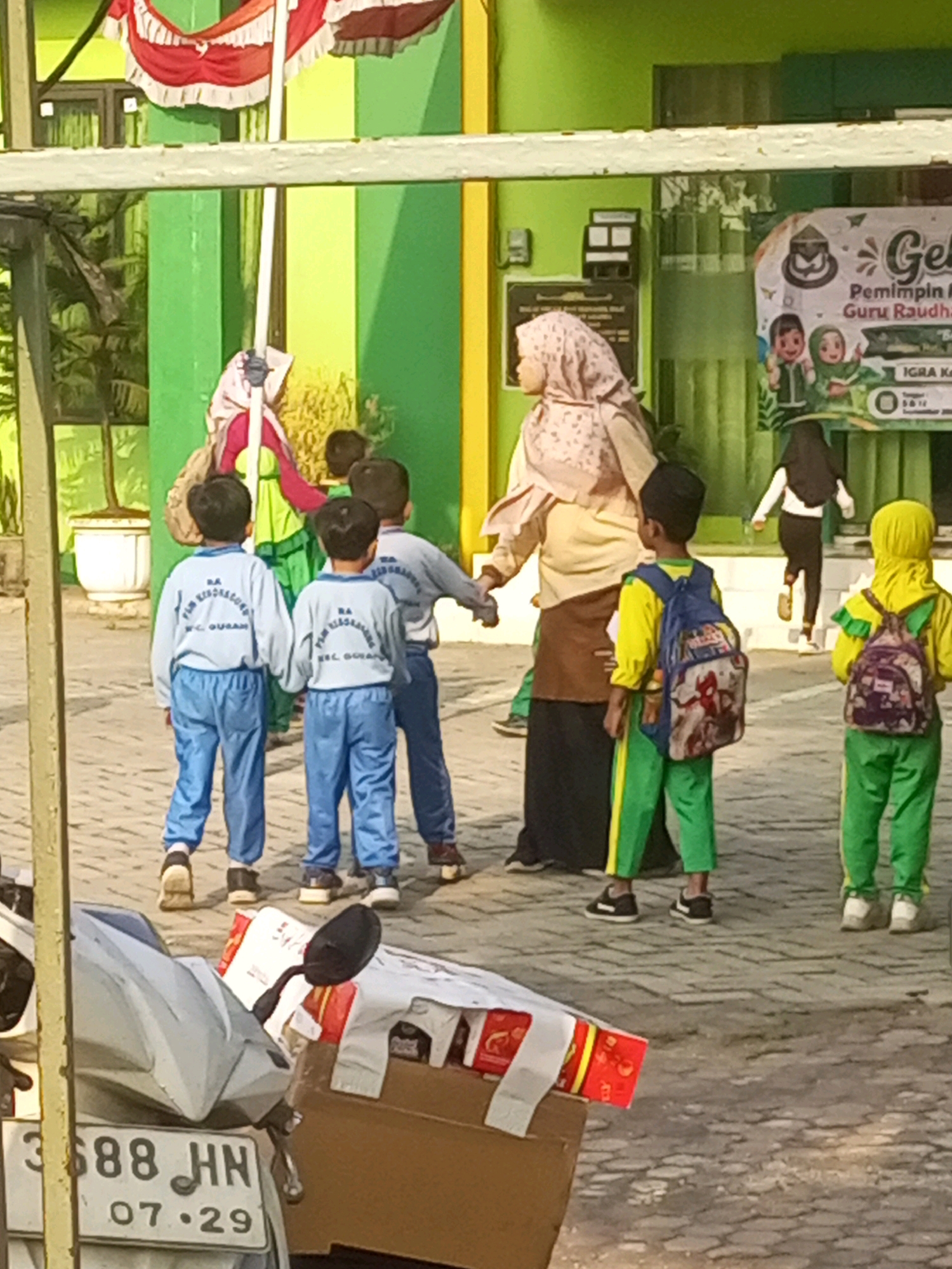 In the city of Gurah, Indonesia, a group of children in uniform are gathered outside a green and yellow building, possibly a school. Several children are wearing light blue shirts with the inscription "RA NURUL BURAH" and matching blue pants. Two children in the foreground are wearing yellow shirts with green pants and backpacks. One backpack features a Spider-Man design. A woman wearing a pink hijab and a light yellow top is standing near the children. In the background, a banner with text and illustrations is visible. The ground is paved with interlocking bricks, and a motorcycle with the license plate "3588 HN 07.29" is parked in the lower left corner of the image. A red and white flag is visible on a pole behind the group.