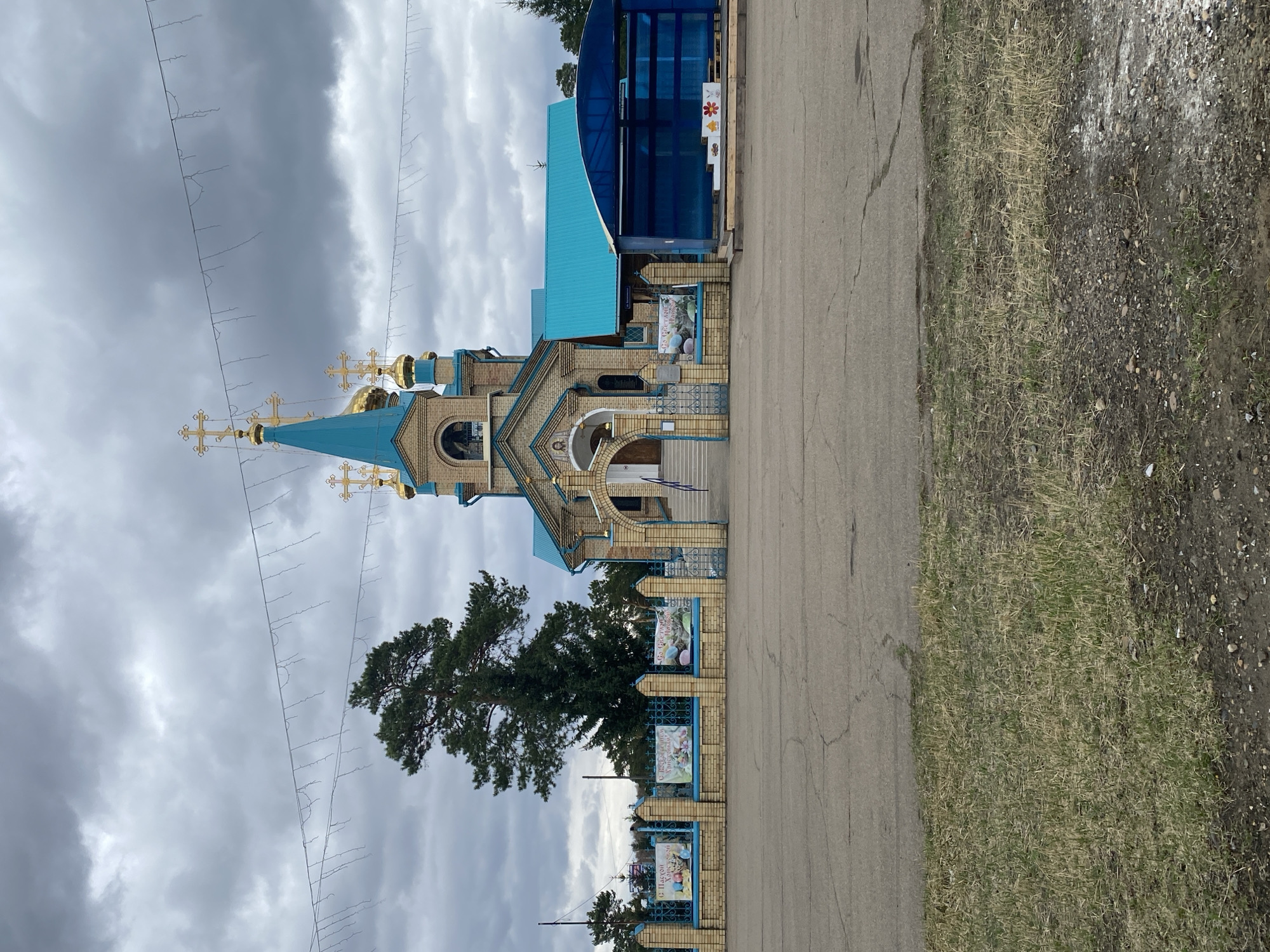 The image shows an Eastern Orthodox church in Sosnovka, Russia. The church features blue roofs and domes, with gold crosses on top. The building is made of light-colored brick and has arched entrances. A brick wall with blue trim surrounds the church grounds, and there are banners attached to the wall. A large tree is located near the church. In front of the church is a paved road, with grass and dirt on the side. The sky is cloudy and overcast. There are string lights hung up in the sky in front of the church. An add-on building is attached to the right side of the main church building.