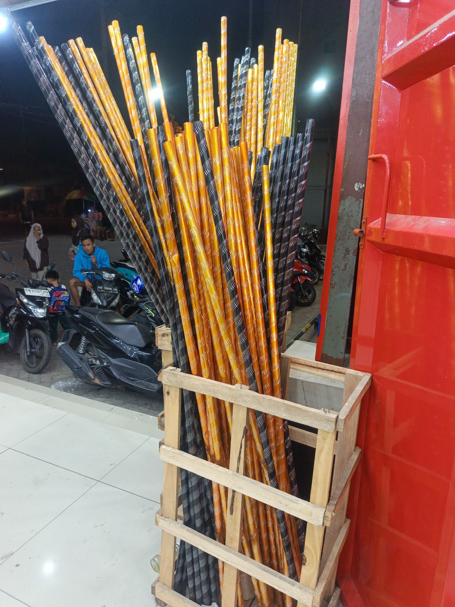 A group of tall, cylindrical objects, appearing to be fireworks or decorative poles, are stored in a wooden crate. These objects are predominantly orange and black with a spiral pattern. The crate is positioned in front of a large, bright red structure, possibly a vehicle or building facade. In the background, several motorcycles are parked. A man in a blue shirt is seated on one of the motorcycles, looking towards the viewer. Another person, a child, is standing near him. In the further background, other individuals and market stalls are visible, suggesting an outdoor commercial area. The lighting indicates it is nighttime or a dimly lit indoor space. The scene is located in Limboto, Indonesia.