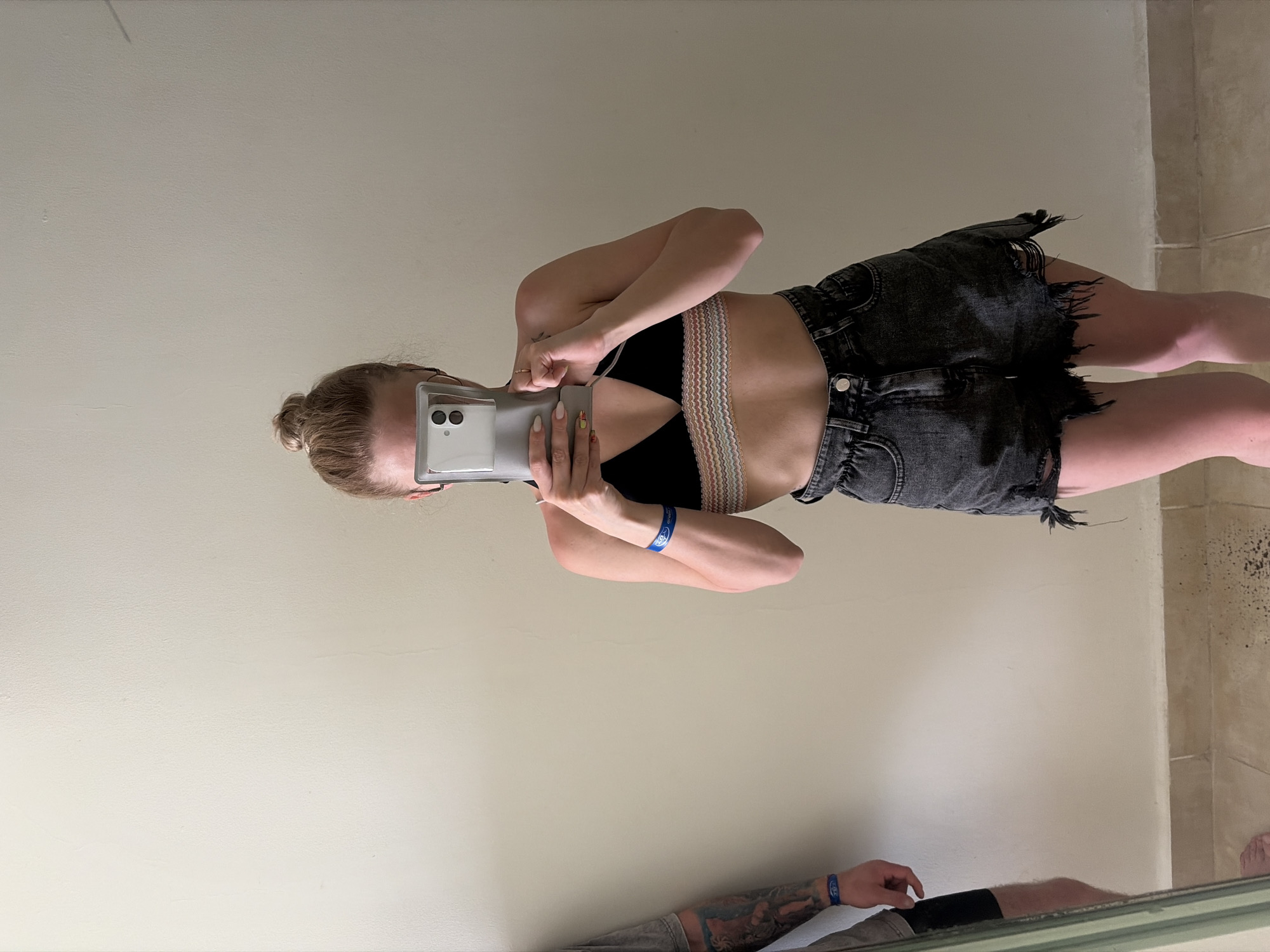 The image is a selfie of a young woman, possibly in her early twenties. She is wearing a black bralette with a multi-colored trim, and distressed dark-wash denim shorts. She is holding a white smartphone in front of her face, obscuring her features. Her hair is pulled back into a bun. She is wearing a blue wristband on her left wrist. The selfie appears to be taken in front of a mirror, reflecting the woman and a portion of a man's body in the lower part of the image. The man has a blue wristband matching the woman's, a tattoo on his arm, and is wearing dark clothing. The setting is inside, likely a bathroom, with plain white walls and a tiled corner visible on the right side of the frame. The location is implied to be Sharm El Sheikh, Egypt, although this detail cannot be confirmed from the image alone.