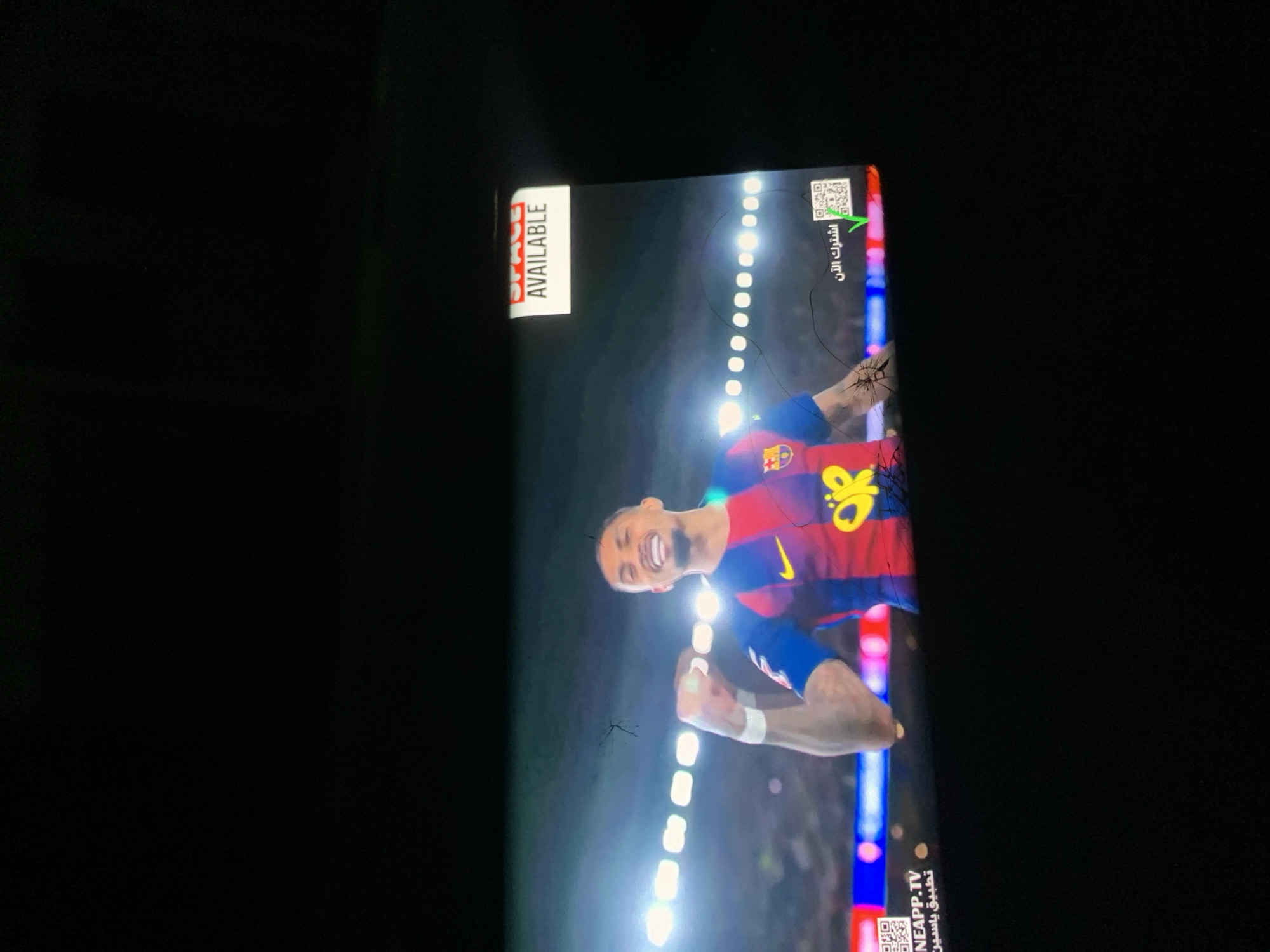 The image shows a screen displaying a football player in a Barcelona jersey, celebrating with his fists clenched and a wide smile. The screen has cracks. Text in Arabic is visible on the right side. A white banner with "AVAILABLE" is affixed to the top left corner. A QR code is visible on the bottom right, next to "NEAPP.TV". The surrounding environment is a dark room. There is no evidence in the image to establish the location as Ungwan Liman, Nigeria.