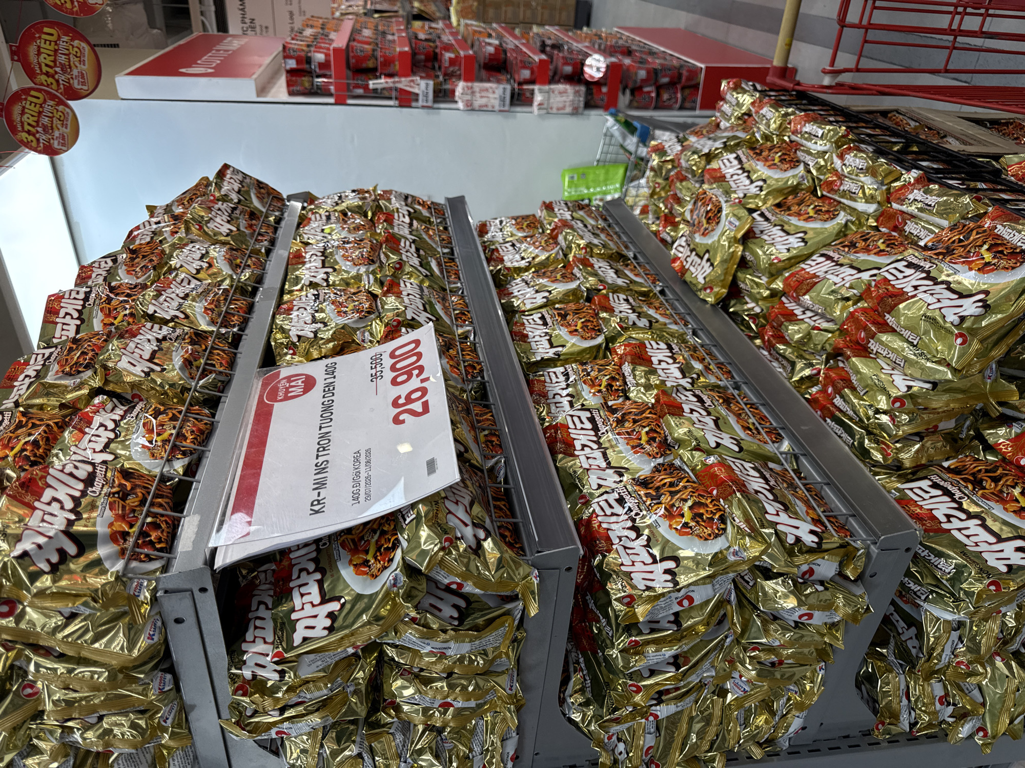 The image shows a retail display of instant noodles, specifically "Chapagetti" brand, at a Lotte Mart store in Nha Trang, Vietnam. The noodles are packaged in shiny gold foil bags with Korean text and images of the prepared dish. The display is organized on metal shelves. A price tag is visible, showing "26,900" in large red digits, with a crossed-out price of "35,500" above it. Red circular promotional signs are hanging from the ceiling, indicating a "3 TRIEU THANH VIEN" (3 million members) campaign. The background shows more retail shelving with other products, and a red shopping cart is partially visible on the right. The lighting suggests an indoor supermarket environment. There are no people clearly visible in the frame.