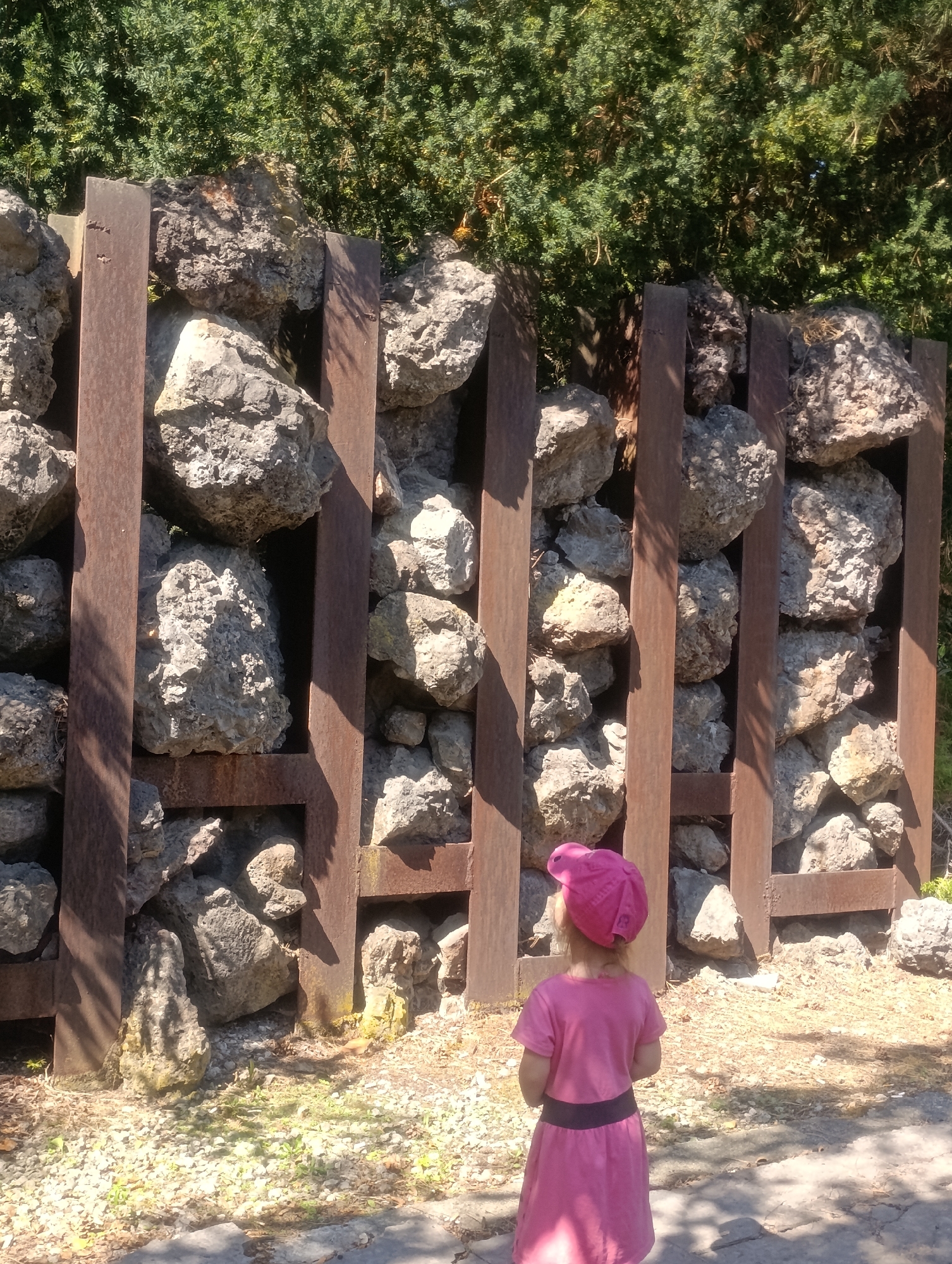 A young child wearing a pink dress and hat stands facing away from the camera, looking at a large structure made of rocks held within a metal frame. The structure is in a park-like setting with green trees in the background and gravel and small plants on the ground. The child is positioned in the foreground, with the rock structure occupying the midground and background. The scene is illuminated by sunlight, casting shadows on the ground. The rock structure appears to be an art installation or a garden feature. This scene is located in the Smetanovy sady park in Olomouc, Czechia.