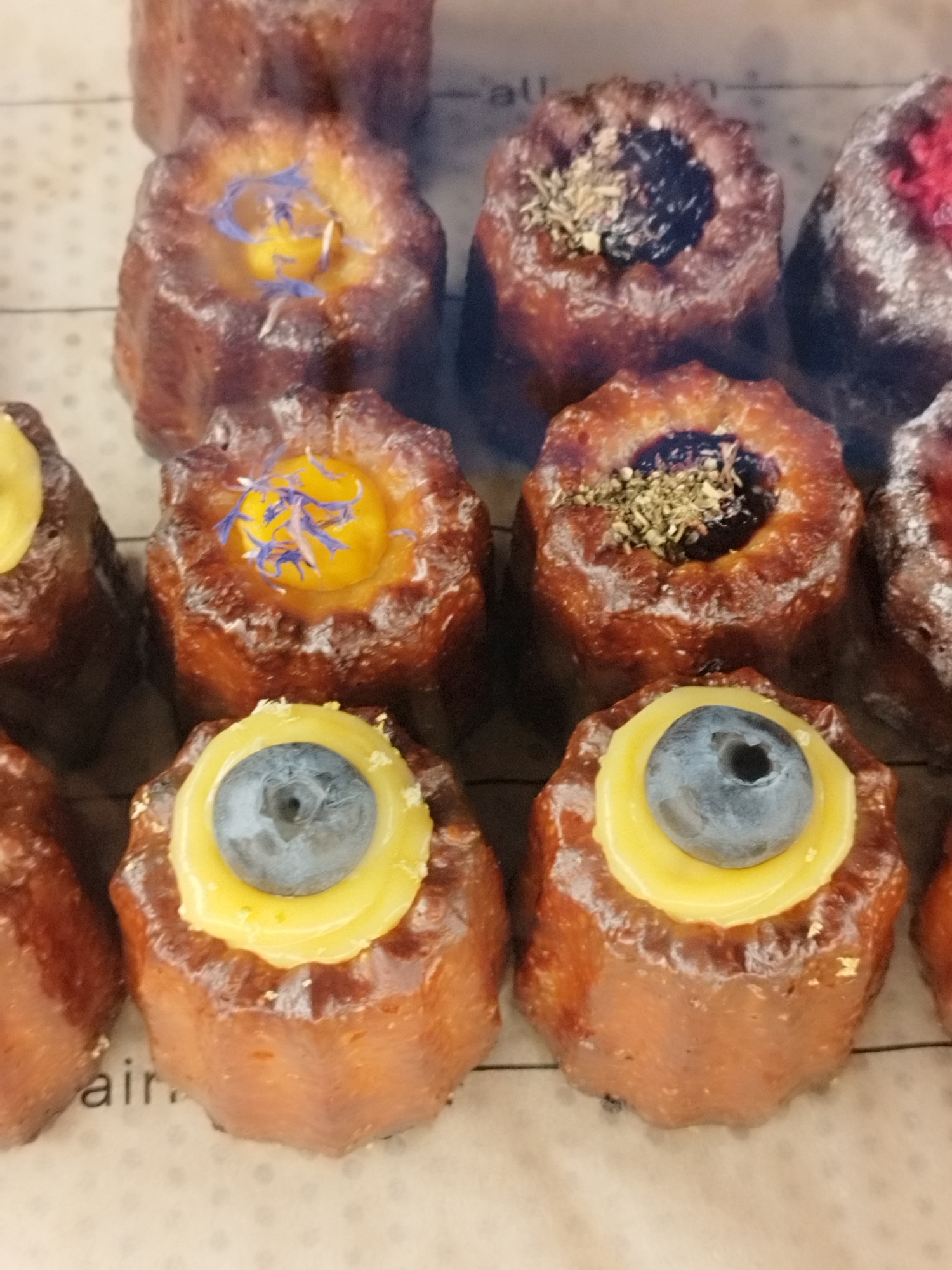 The image displays a close-up view of several canelés, a French pastry, arranged in a grid. The canelés are small, cylindrical, and have a dark, caramelized crust. They are topped with various garnishes. Some are decorated with a dollop of yellow cream and blue cornflower petals, while others have a dark filling topped with dried herbs. One canelé in the lower right appears to have a red topping, possibly berries.
The canelés are presented on a light-colored surface with a subtle dotted pattern. In the background, there is a hint of text on a light surface, with the letters "air" clearly visible at the bottom left of the frame. The lighting is bright, suggesting an indoor setting, likely a bakery or patisserie. The overall impression is one of artisanal desserts, emphasizing texture and color. The specific location cues, such as the text "air" and the implied setting of a bakery, are consistent with a context in Saint Petersburg, Russia. There are no people visible in the image. The focus is entirely on the pastries themselves, showcasing their different toppings and textures.