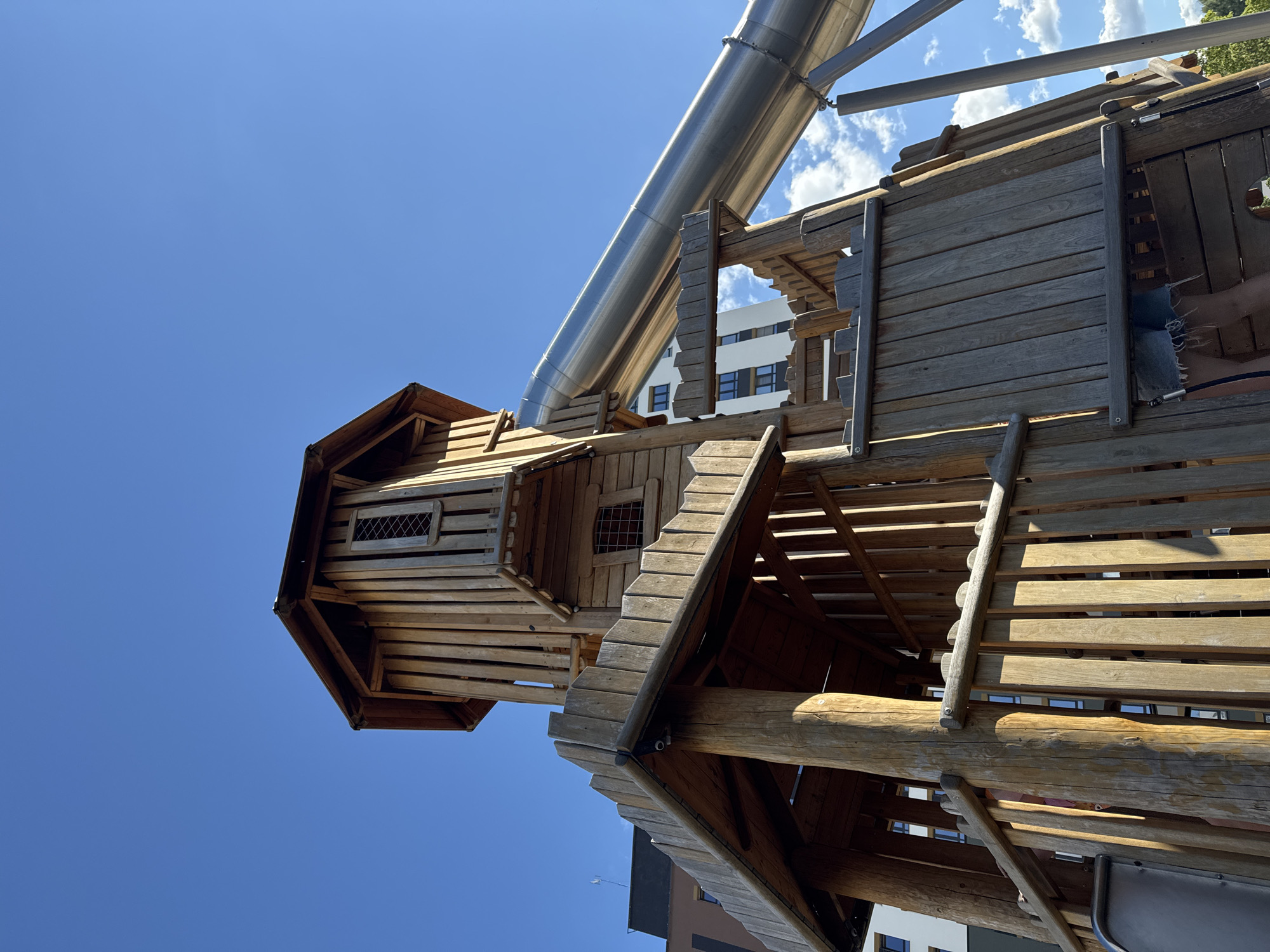 This image features a wooden playground structure with a large, silver slide. The structure appears to be a multi-level playhouse or fort, built with logs and wooden planks, some of which show signs of wear. A silver tube slide angles downwards from a higher platform. In the background, a modern white building with dark windows is visible, suggesting an urban or semi-urban setting. The sky is bright blue with scattered white clouds, indicating it is daytime and likely sunny. The perspective is from below, looking up at the play structure and the sky. There is no visible text in the image. The scene is set in Patrusheva, Russia.