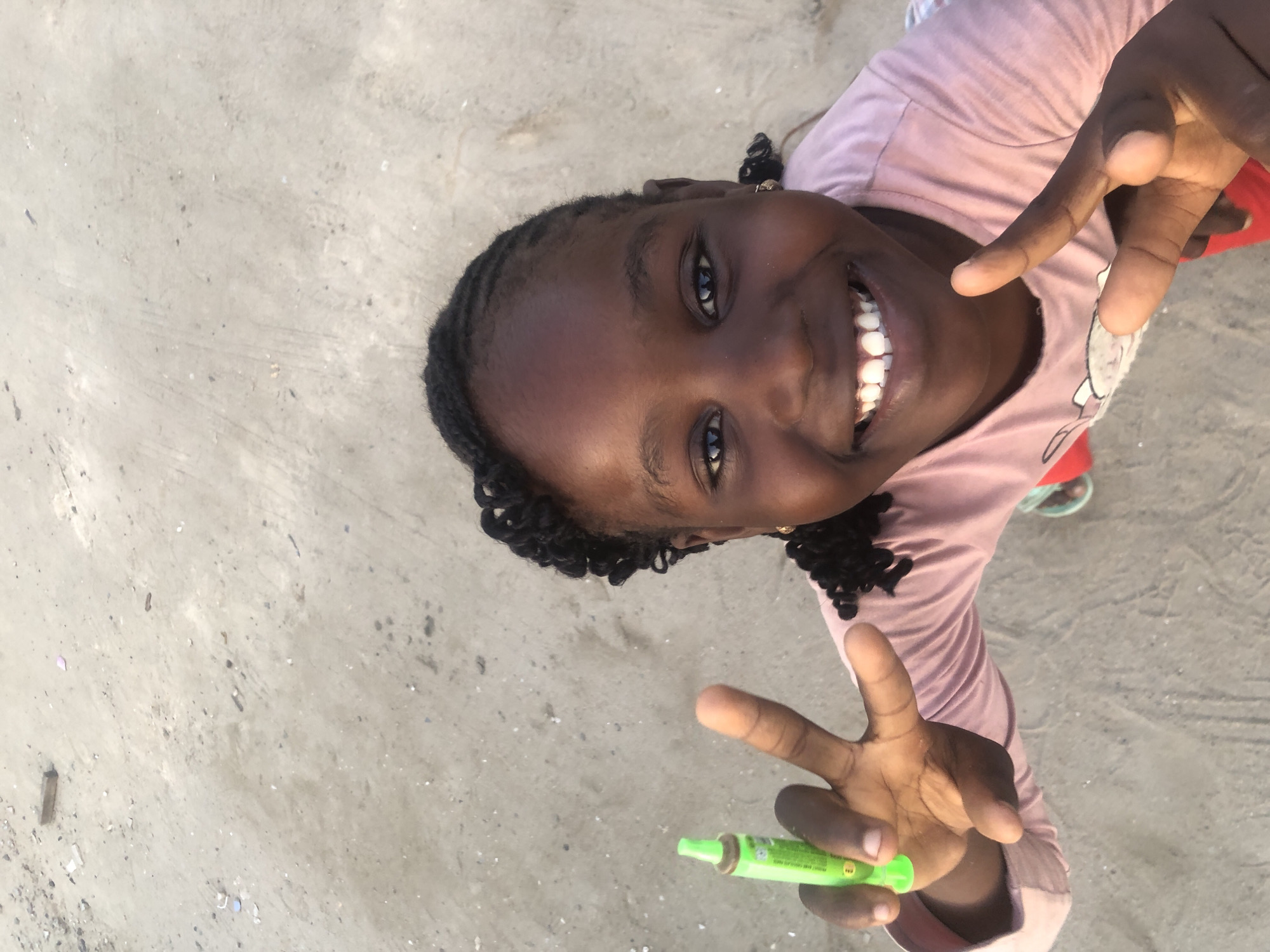 An overhead, medium shot captures a young Black girl with braided hair smiling widely and holding up two fingers in a peace sign. She is wearing a pink t-shirt and red shorts. In her left hand, she holds a small, green tube. The ground beneath her is sandy and appears to be a public space within the city of Ado, Nigeria. The scene is outdoors and appears to be during daylight.