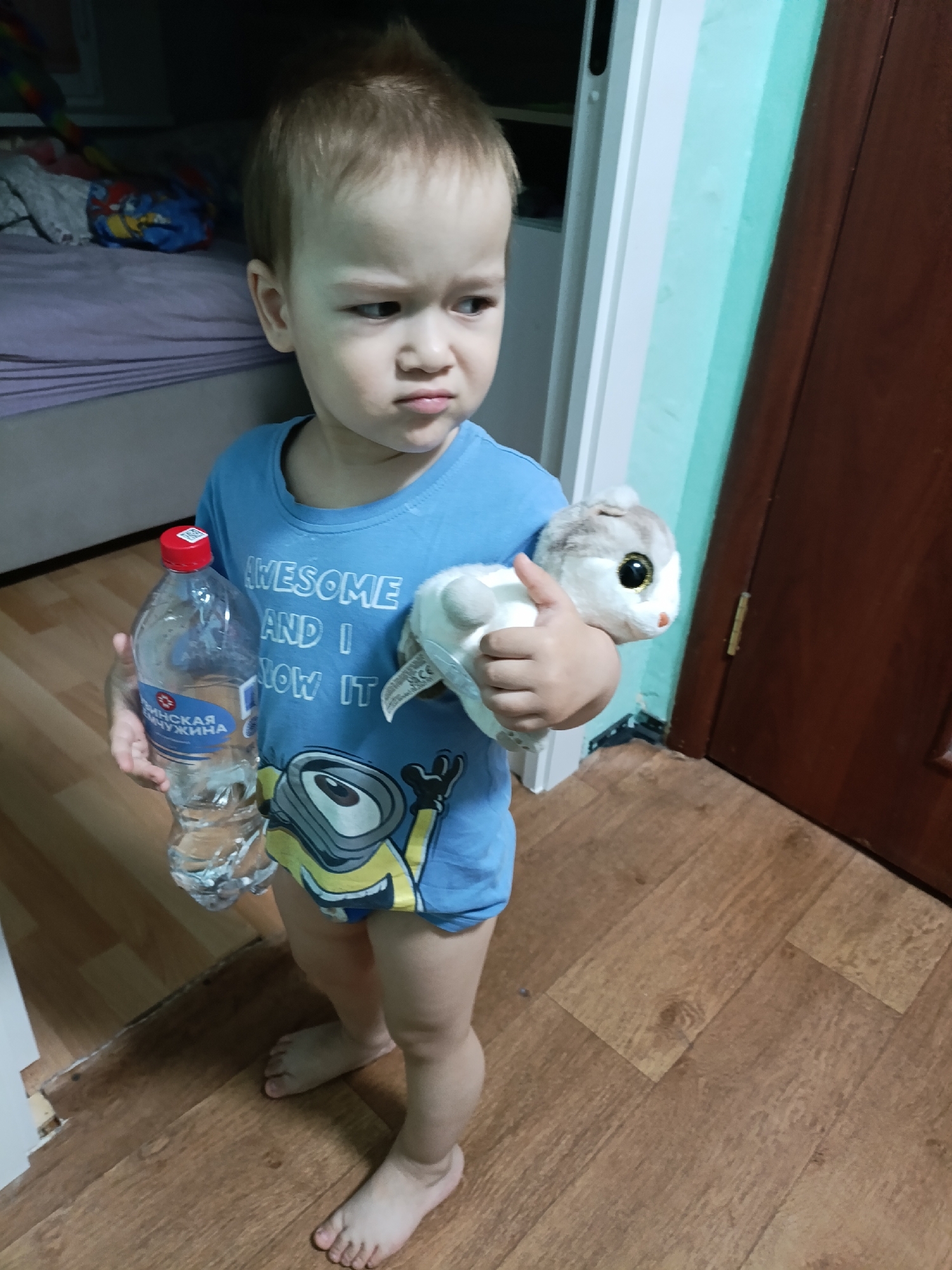 A young child stands indoors on a wooden floor, holding a water bottle and a stuffed animal. The child is wearing a blue t-shirt with a Minion character on it. The background includes a bed with bedding, a door frame, and part of a wooden door. The water bottle label includes Cyrillic text and indicates "Чайковский" which may reference the city of Chaikovsky, Russia. The child's expression is pensive.