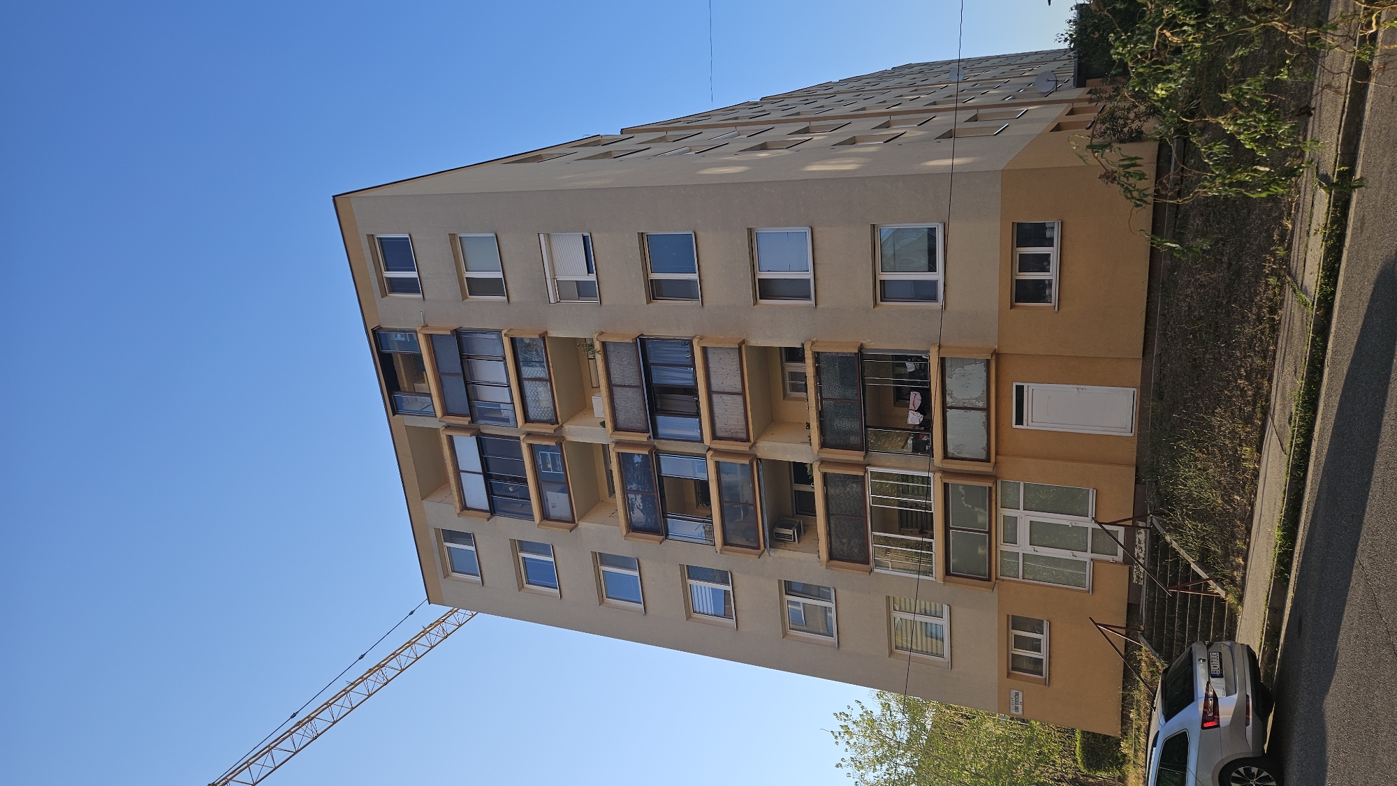 An exterior, eye-level, long shot captures a multi-story apartment building and a construction crane under a clear blue sky. The building, constructed of concrete and painted beige and yellow, features multiple balconies with glass enclosures. A silver station wagon is parked on the street in front of the building. The building is located at 3054 Vlčkova Street in Bratislava, Slovakia.