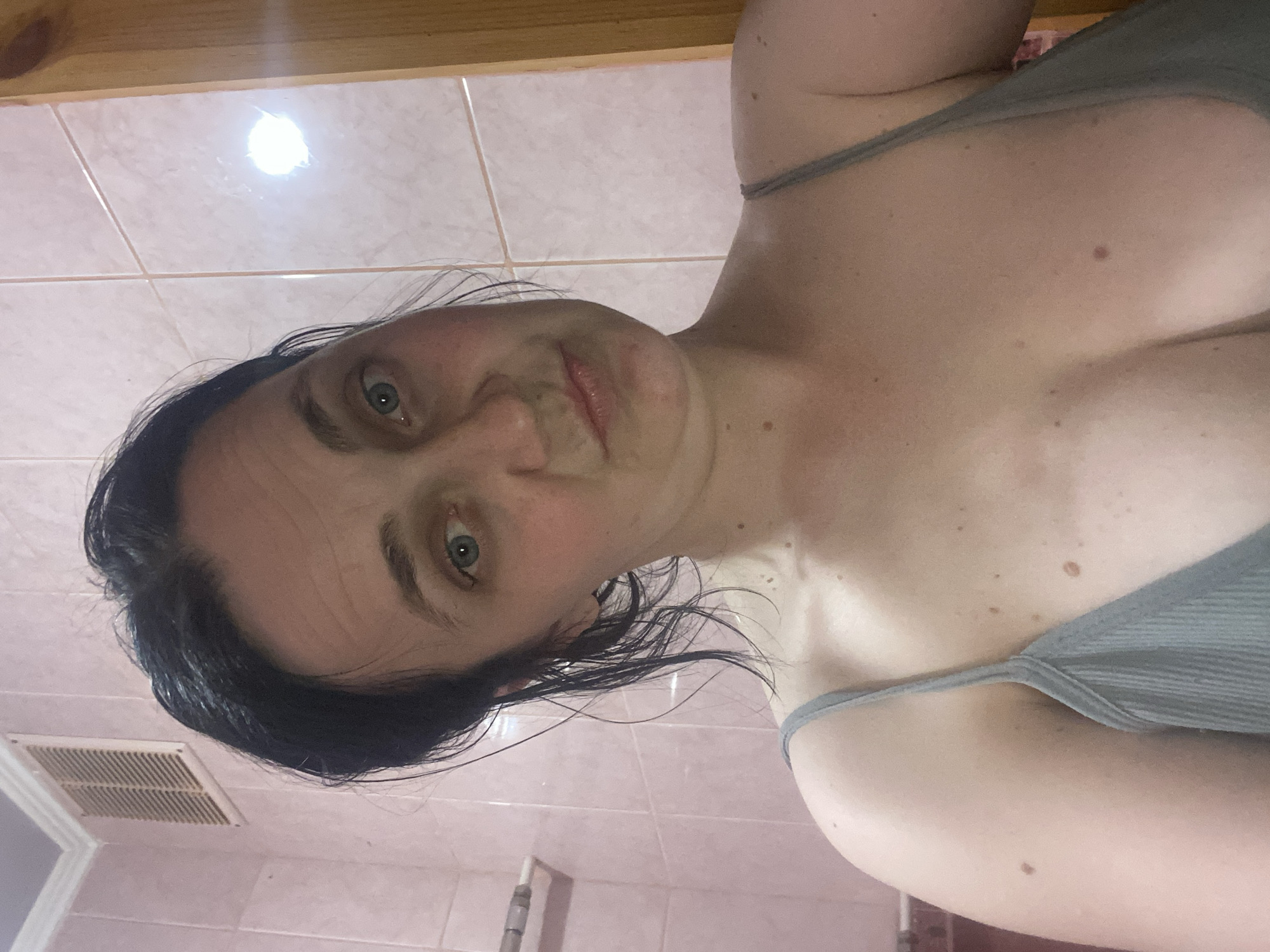 A young woman with dark hair and green eyes is taking a selfie in what appears to be a bathroom. She is wearing a light gray camisole. The background consists of pink tiles and a wooden door frame. A ventilation grate is visible in the upper left corner.