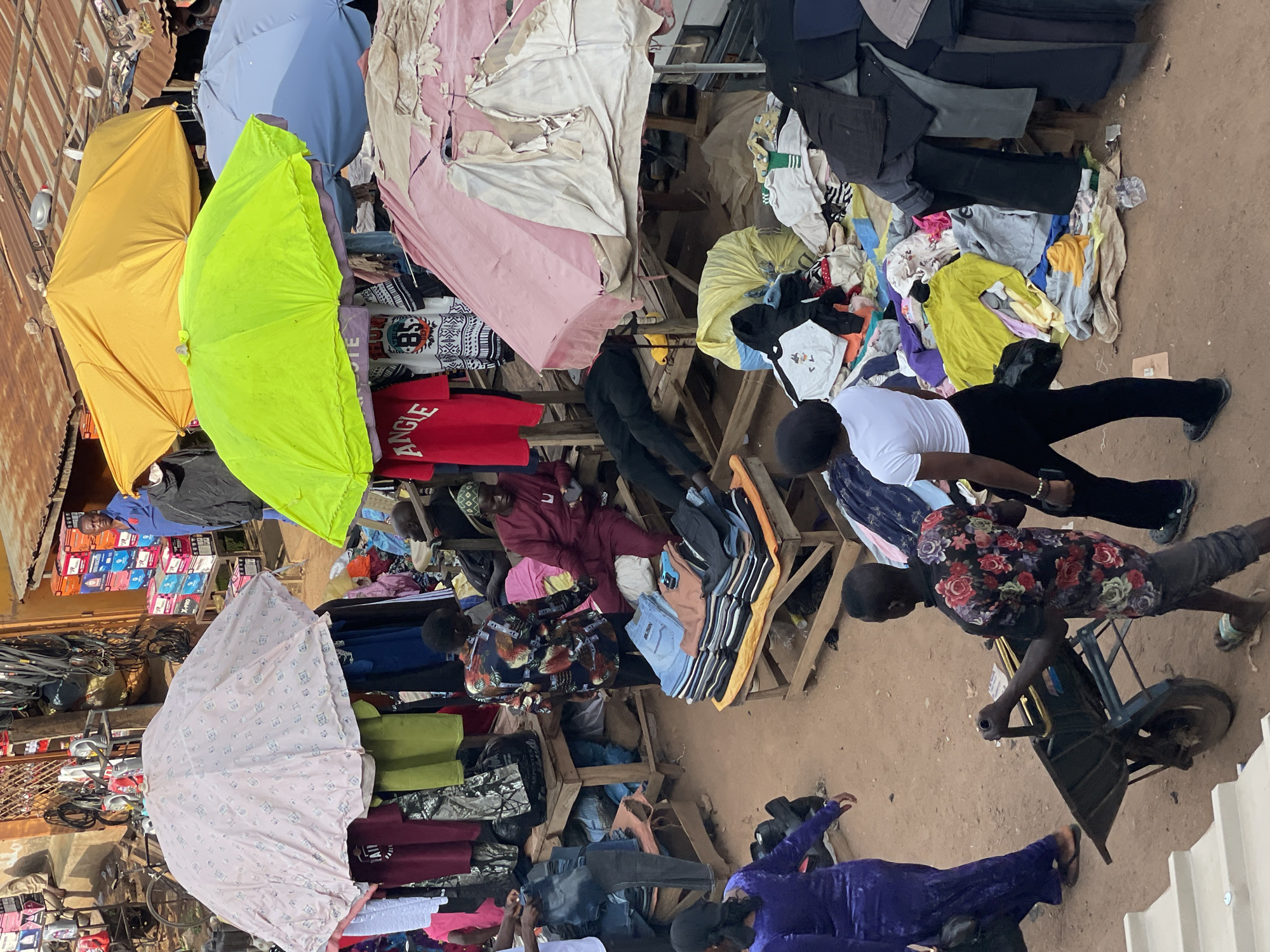 The image captures a bustling scene at an open-air market in an unidentified city. Several vendor stalls are visible, each displaying a variety of clothing items ranging from t-shirts and jeans to dresses. These stalls are shaded by umbrellas of various colors, including yellow, green, blue, and pink. Multiple people are present, browsing through the clothing selections. One person is operating a wheelbarrow in the foreground. The ground appears to be dirt.