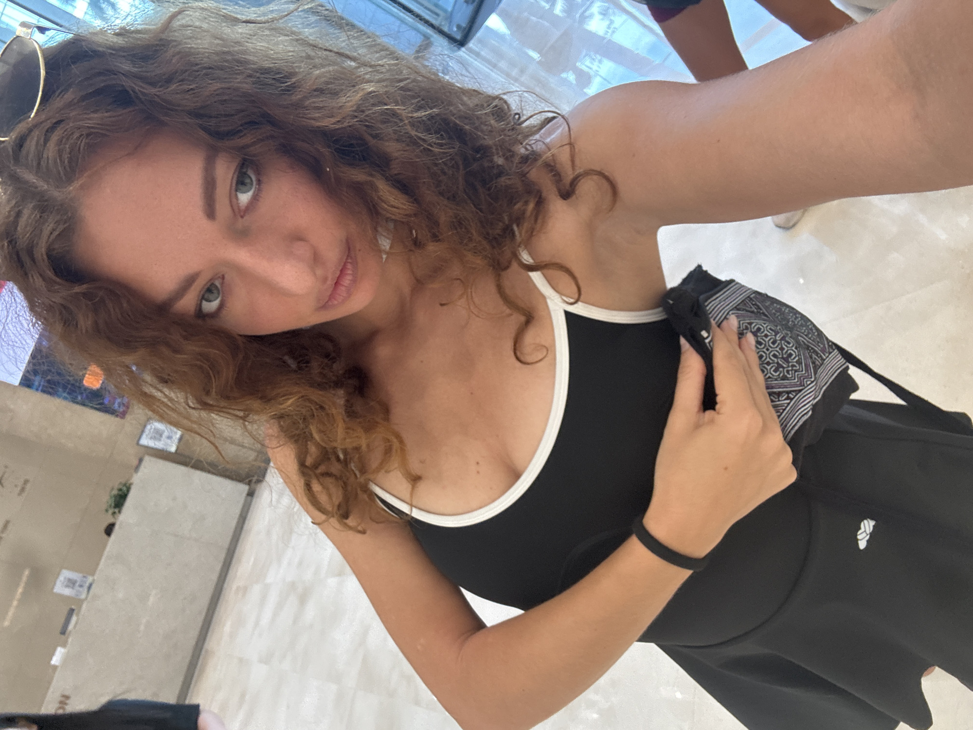 A woman with long, curly, reddish-brown hair is taking a selfie in an indoor public space. She is wearing a black sleeveless top with a white trim and a black skirt. Her left hand is holding a black patterned cloth to her chest, and she has a black bracelet on her left wrist. Sunglasses are perched on top of her head. The background shows a reception desk and a tiled floor. The lighting suggests it is daytime. The location is identified as Hanoi, Vietnam.