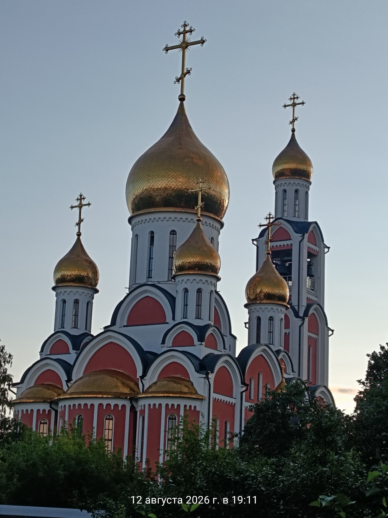A close-up, low-angle shot captures the golden domes and red and white architecture of a Russian Orthodox church against a clear, pale blue sky. The church features multiple domes of varying sizes, each topped with a cross. A prominent bell tower is visible on the right side of the image. Green foliage frames the lower portion of the structure, with some trees obscuring the base. The image is dated "12 августа 2026 г. в 19:11" in the lower right corner.