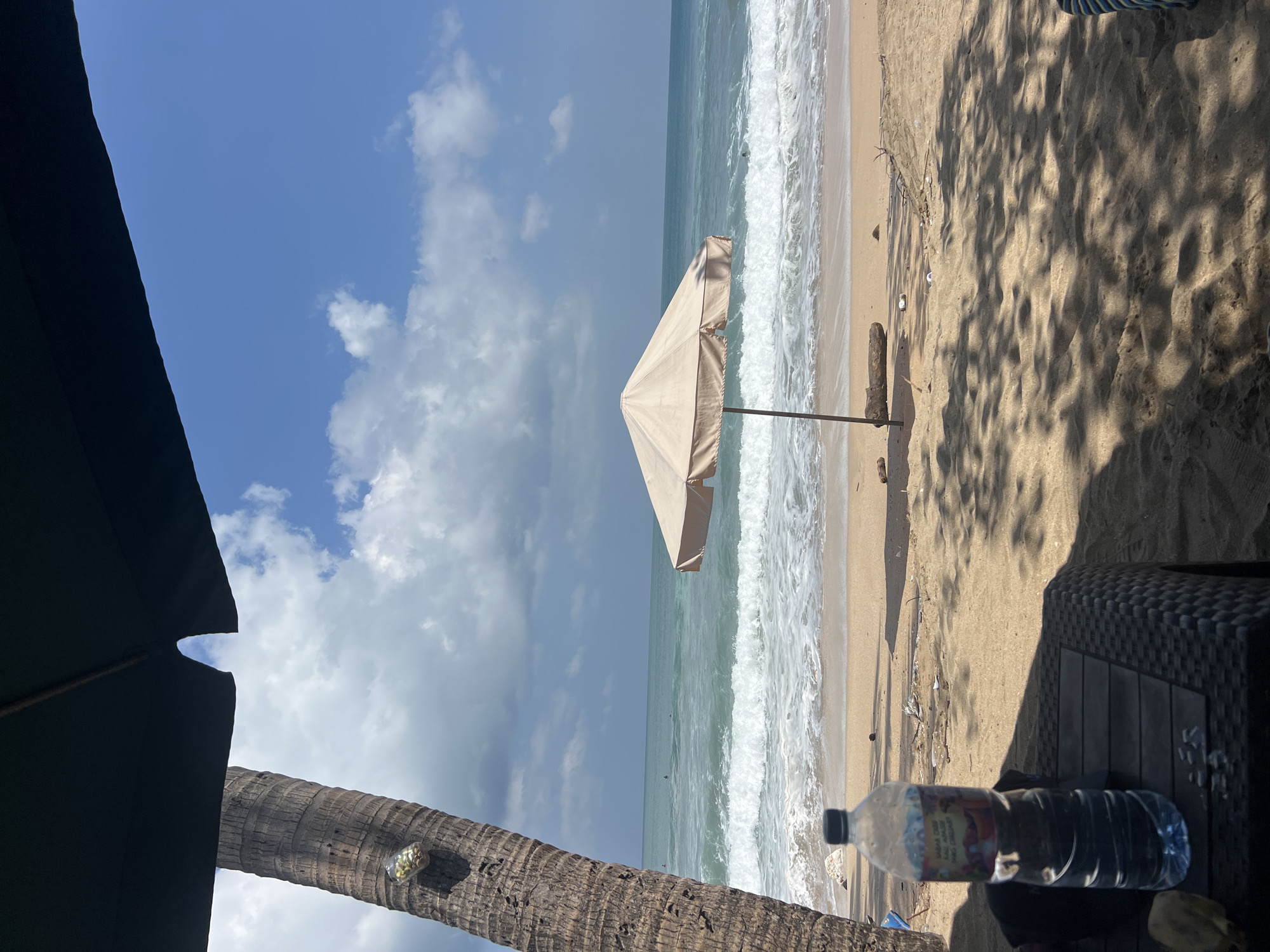 The image depicts a tranquil beach scene in Kuta, Indonesia, under a clear blue sky with scattered white clouds. A palm tree stands to the left, partially shaded by a dark umbrella. A light-colored beach umbrella is positioned in the sand near the water's edge. The ocean waves are breaking on the shore, and a few individuals can be seen in the water. In the foreground, a plastic bottle of water rests on a dark, textured surface, likely a beach chair. The sand is adorned with shadows from the palm tree and umbrella, creating a dappled effect. Scattered debris, including small white containers and dried vegetation, is visible on the sand. The overall atmosphere is one of peaceful relaxation.