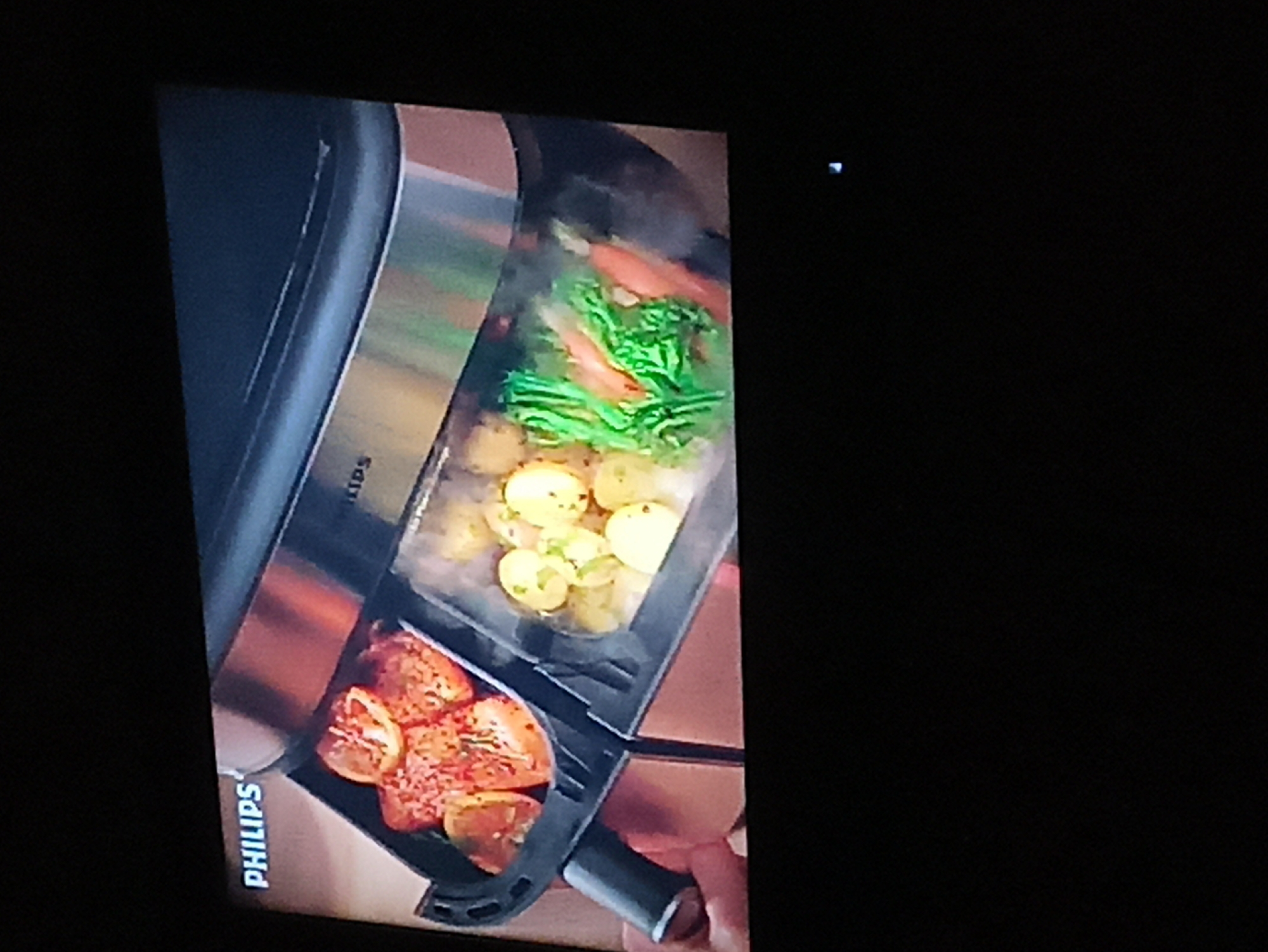 The image shows a close-up of a Philips air fryer with food inside. The air fryer appears to be new and clean, with a sleek design. In the bottom compartment, there are sliced oranges. In the main section, there are potatoes, green vegetables and salmon filets. Steam can be seen rising from the vegetables. The background is dark.