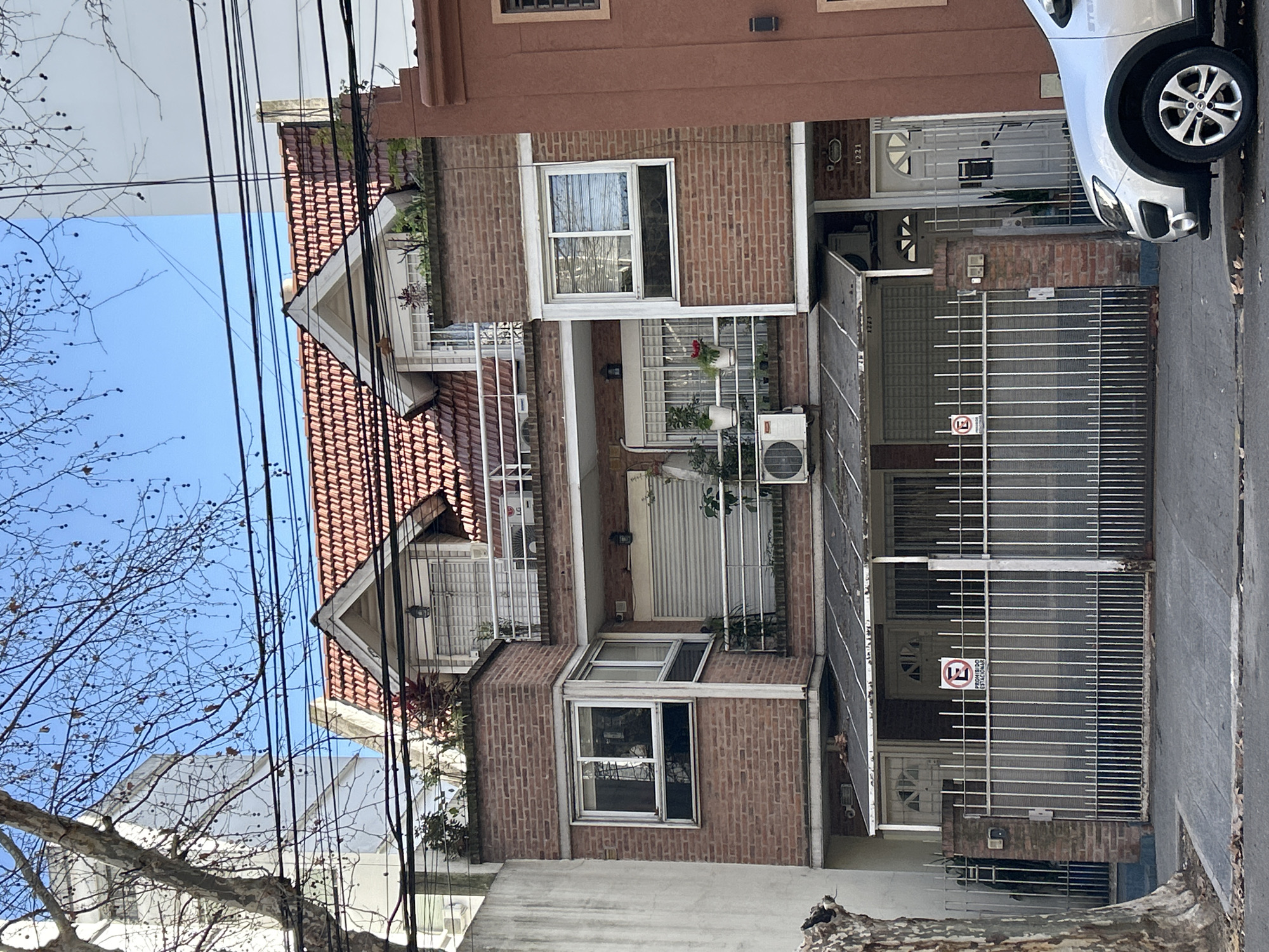 The image depicts the facade of a multi-story brick building on a city street, likely in Buenos Aires, Argentina, judging by the architecture and the "Prohibido Estacionar" sign visible on the gate. The building has a red tile roof with dormer windows. The lower level features a metal gate providing access to two garage entrances, one of which has a "Prohibido Estacionar" sign. A silver car is parked on the street in front of the building. The street is paved, and fallen leaves are scattered on the ground. Overhead, electrical wires are strung across the scene. The building exhibits multiple windows, balconies, and air conditioning units. The architecture suggests a residential or mixed-use building.