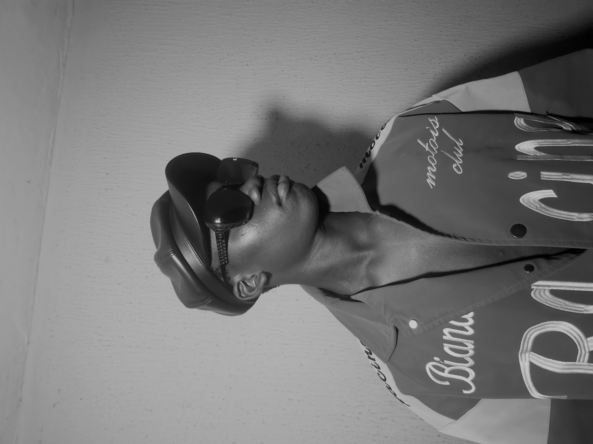 A person wearing a cap and sunglasses is photographed indoors against a textured wall. The individual is wearing a jacket with text embroidered on it. The text includes "motois club" and "Bian." The image is in black and white. No information is present to confirm its location in Nkwelle, Nigeria.