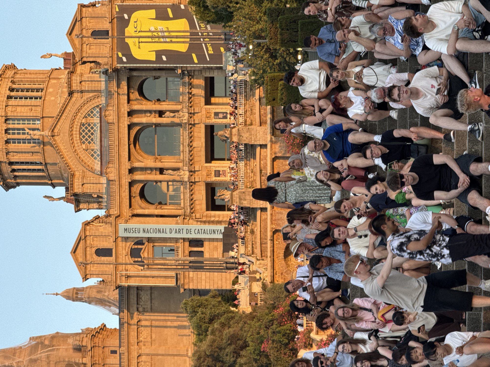 This is an eye-level medium shot of a crowd of people sitting on stone steps in front of the Palau Nacional in Barcelona, Spain. The Palau Nacional is a large, ornate building with a dome and columns, bathed in warm, late afternoon sunlight. A large yellow banner depicting a cycling jersey, possibly from the Tour de France, hangs on the right side of the building, alongside a vertical banner that reads "MUSEU NACIONAL D'ART DE CATALUNYA". The crowd is diverse in age and ethnicity, with many individuals looking at their phones. Some people are interacting, like the two young men in the foreground who appear to be having a conversation. Others are seated alone or in small groups, some appearing relaxed, others engaged in conversation. The steps are covered in people, creating a sense of a communal gathering. Lush green trees and flowering bushes are visible to the left of the steps, adding a natural element to the urban landscape. The sky is a clear, pale blue.