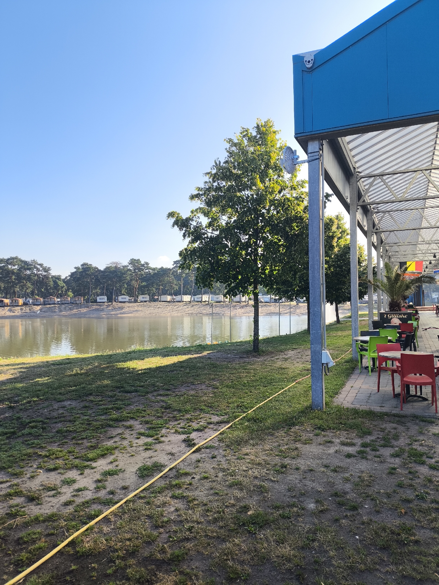 This media file captures a serene waterside scene at a recreational area, likely a beach or lake resort, under a clear, bright sky, indicating it's daytime with good weather. The foreground features a patch of uneven, grassy ground with some bare soil, bisected by a long, yellow hose. To the right, a modern, open-air structure with a blue roof and a white, corrugated ceiling provides shade over a paved patio. This area is furnished with several small, round tables and chairs in vibrant colors: red, green, and black. Some of these tables appear set for patrons, suggesting a cafe or restaurant is in operation.
Beyond the patio, a calm body of water reflects the surrounding environment. Along the far bank of the water, a sandy strip is lined with numerous mobile homes or caravans, and a dense line of tall, green trees forms the backdrop. The trees closest to the water exhibit sunlight dappling through their leaves, while a larger tree stands prominently in the middle ground. The overall atmosphere is peaceful and invites relaxation.
Notable details include a security camera mounted on the blue facade of the structure. In the distance, blurred figures of people are visible, either relaxing on the beach or seated at the tables. A palm tree adds a touch of tropical flair. Text is visible on a black sign beneath the palm tree, reading "t Gilliske". The scene's lighting suggests it could be morning or late afternoon, given the angle of the sun casting long shadows. The location is identified as Lommel, Belgium, which aligns with the presence of such recreational facilities and the European style of the caravans.